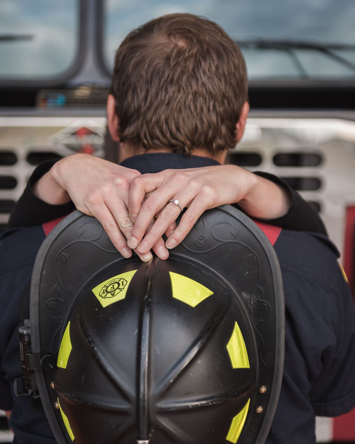 Close up of hands with wedding ring resting on black firefighter helmet against fire truck.