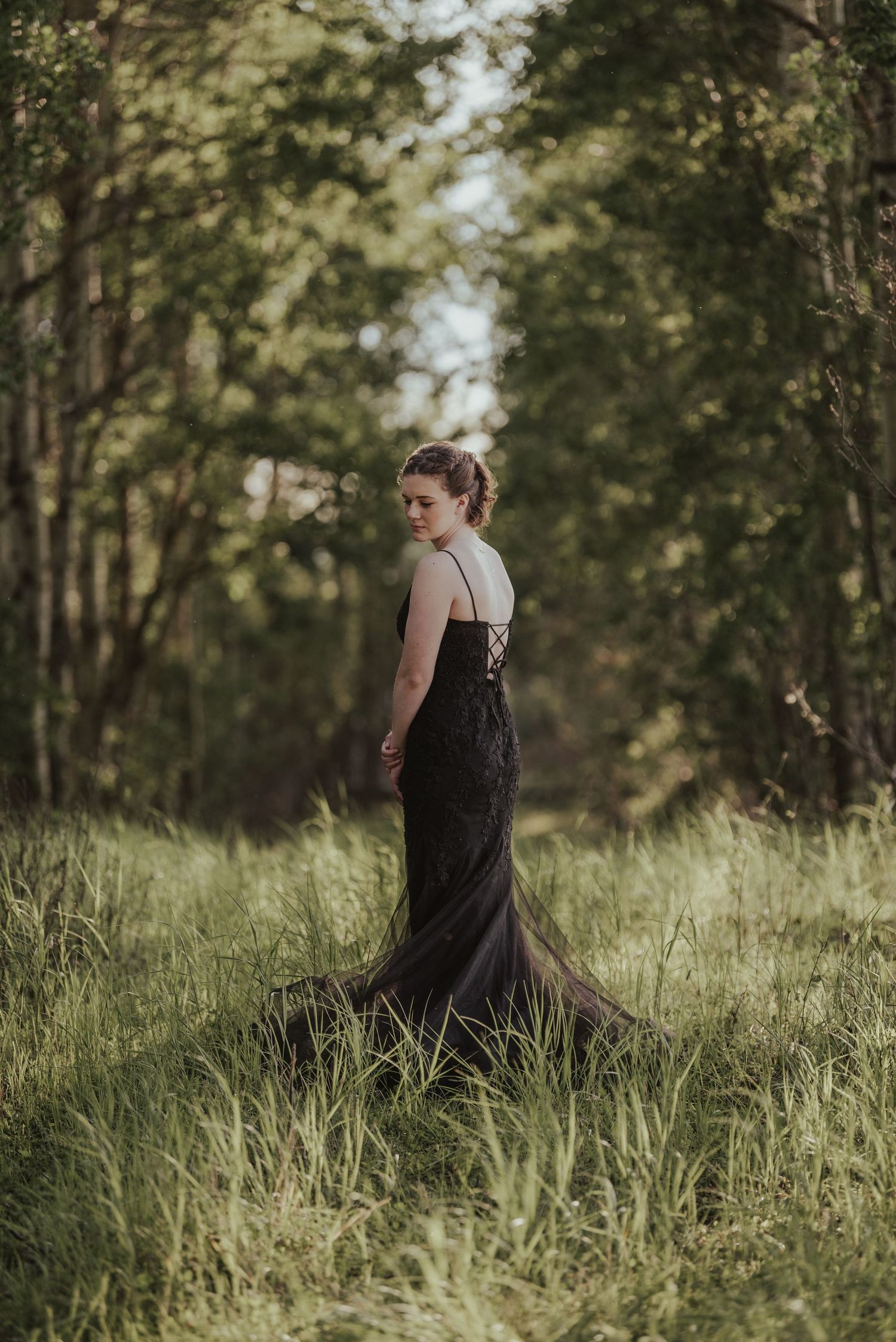 Woman in elegant black gown walking through sunlit forest clearing.