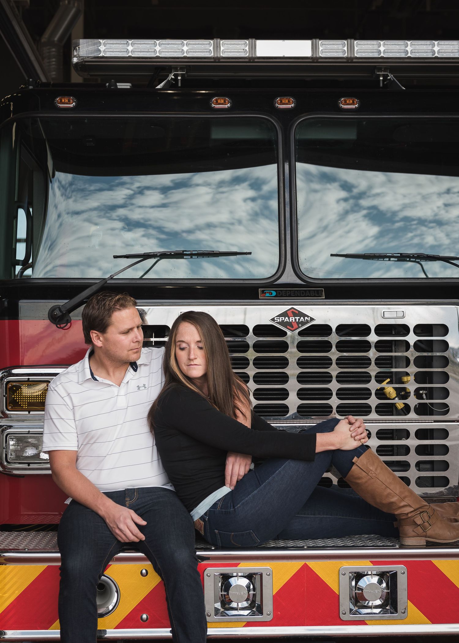 Romantic couple sitting on the front bumper of a red fire truck against a cloudy sky background.