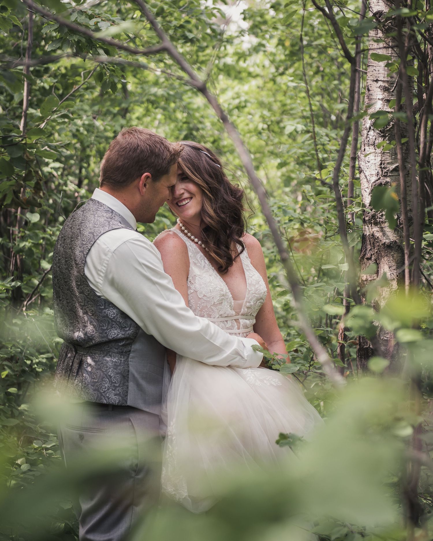 Romantic wedding portrait in birch forest with couple embracing among green leaves and natural light.