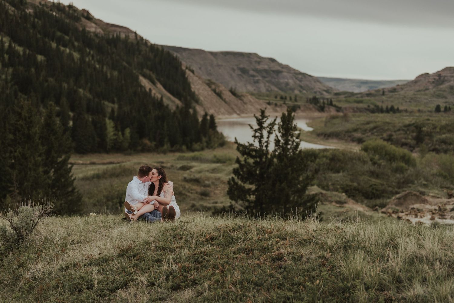 Engagement photo of couple sitting on grassy hillside overlooking scenic mountain valley.