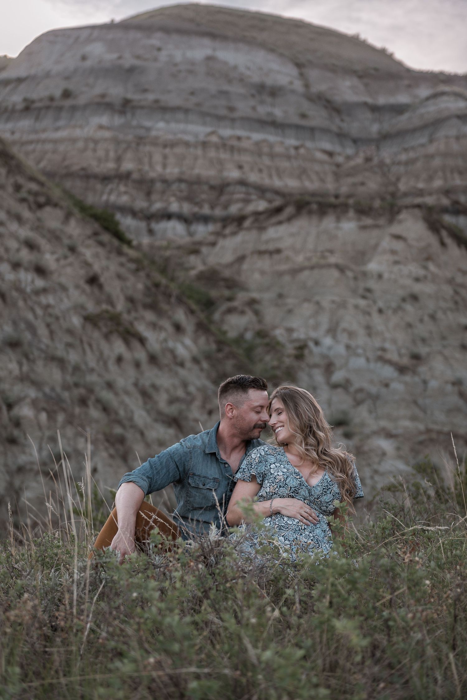 A romantic couple sits together in tall grass with dramatic rocky cliffs and layered badlands formations in the background.