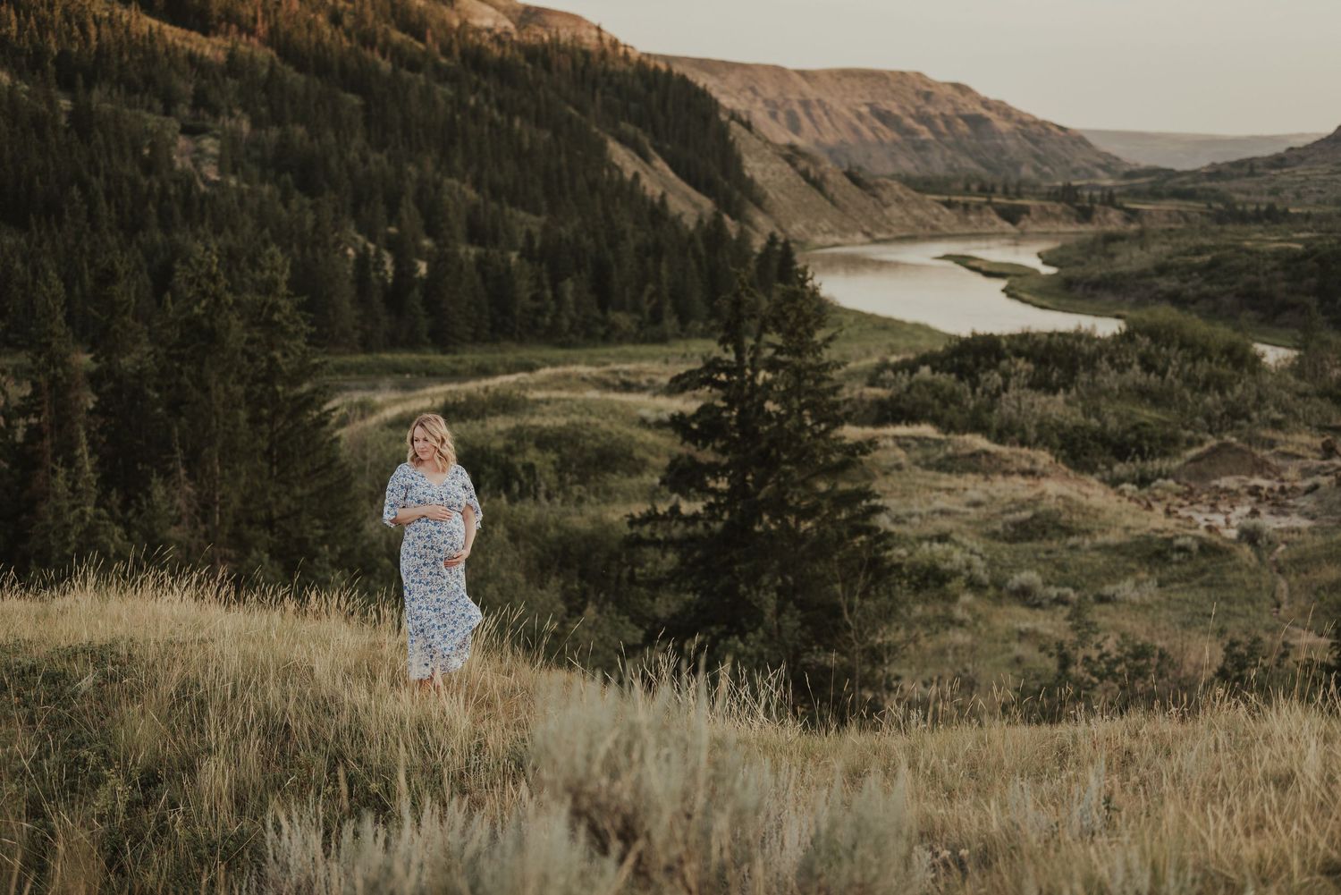 Person in flowing dress stands on hillside overlooking dramatic river valley landscape.