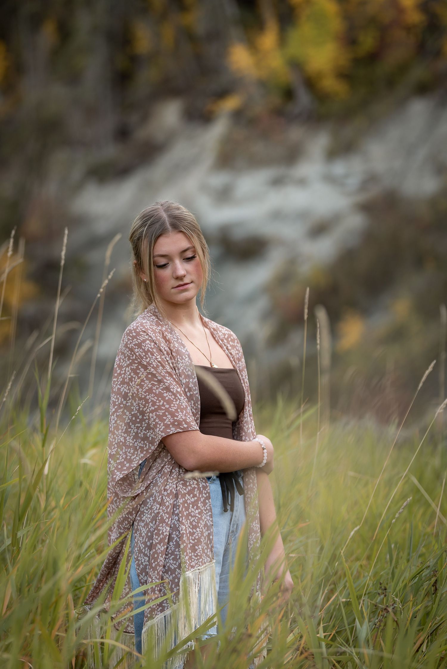 Person in floral shawl standing thoughtfully in tall grass against rocky backdrop.