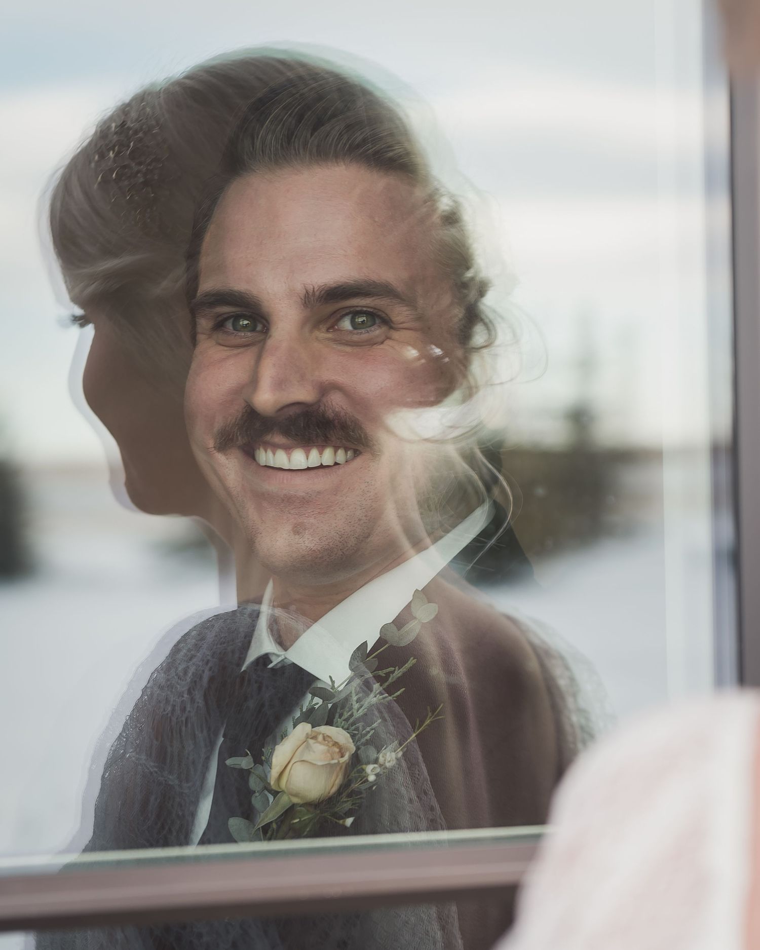 Artistic double exposure portrait of groom smiling with boutonniere through window reflection.