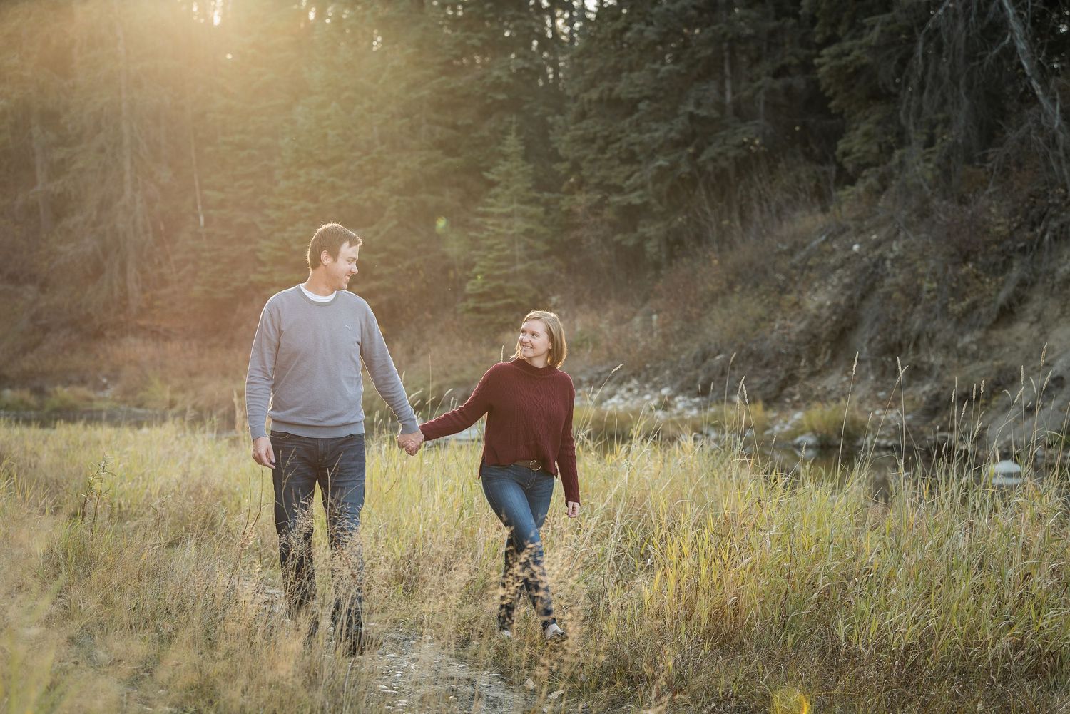 A couple walks hand in hand through a sunny meadow with evergreen trees in the background at golden hour.