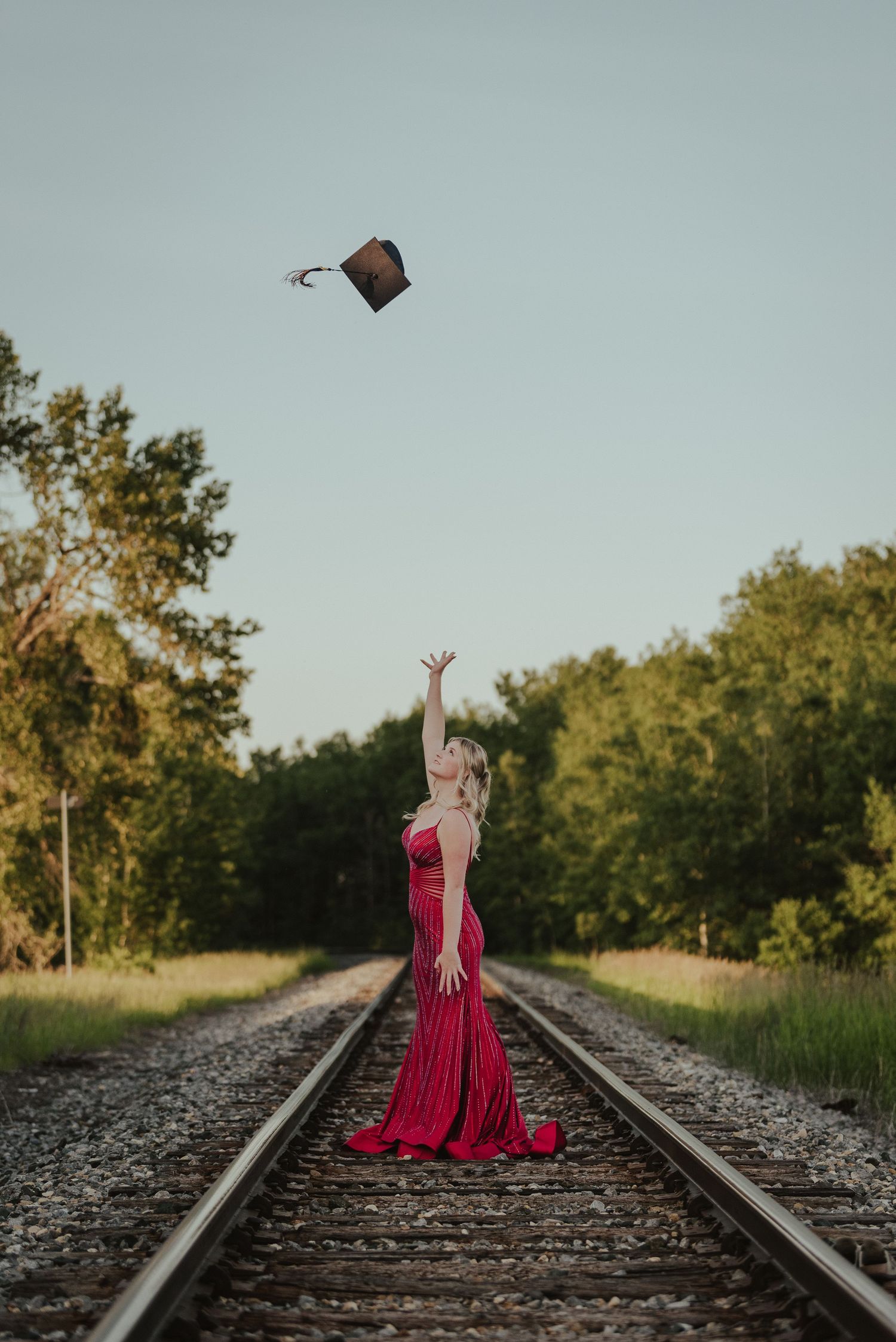 Person in a flowing red dress throwing a graduation cap while standing on train tracks at sunset.