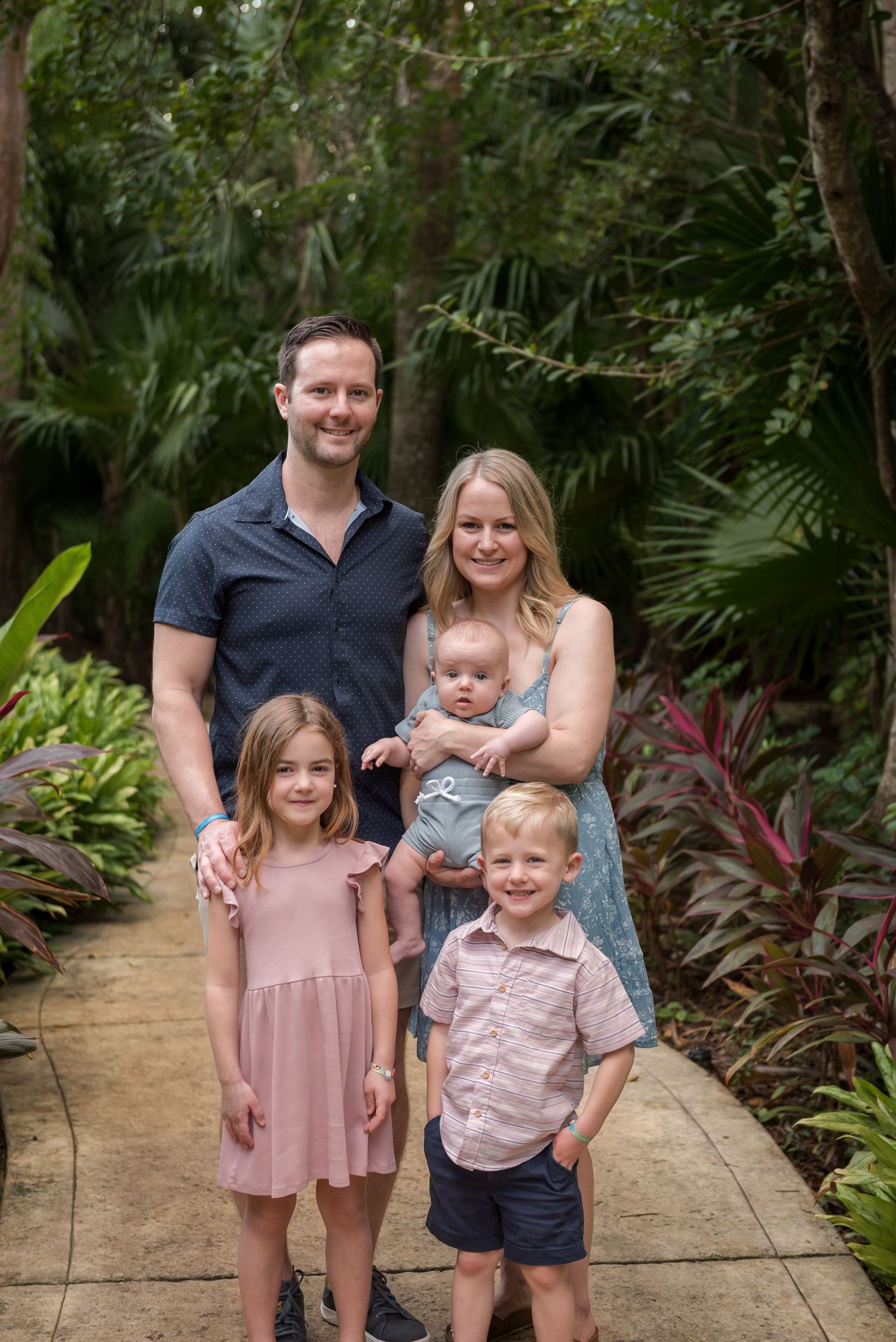 A family of five poses together on a paved path surrounded by lush tropical garden foliage and palm trees.