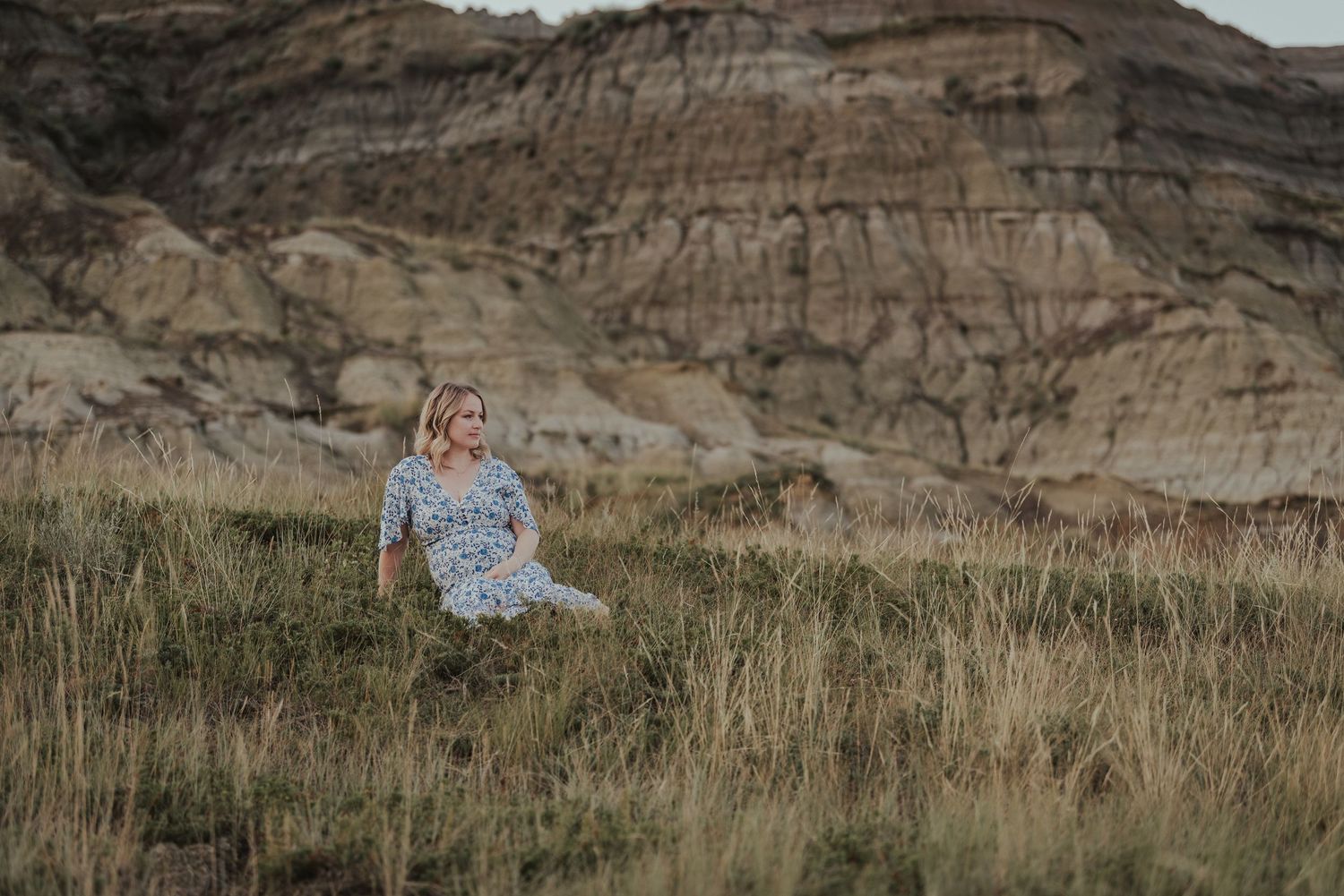 Person in a dress sitting in tall grass with rugged badlands formations in the background at sunset.