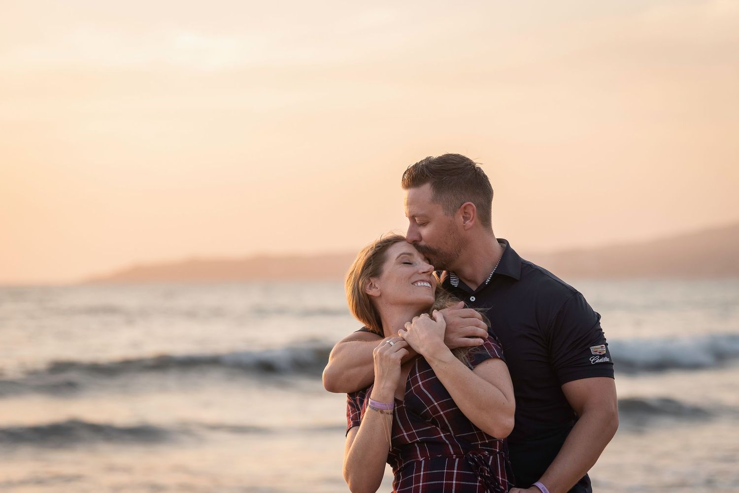 A couple sharing an intimate moment at sunset on the beach as waves roll in behind them.