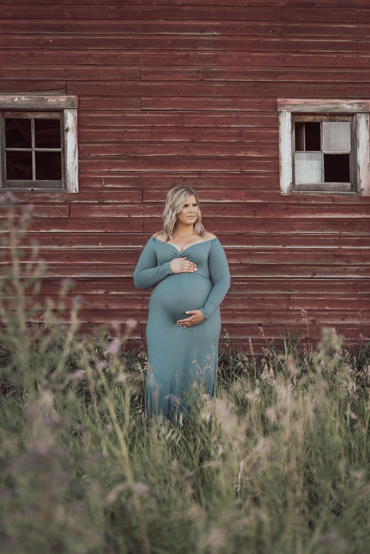 An expectant mother in a teal dress stands in tall grass before a rustic red barn with weathered windows.
