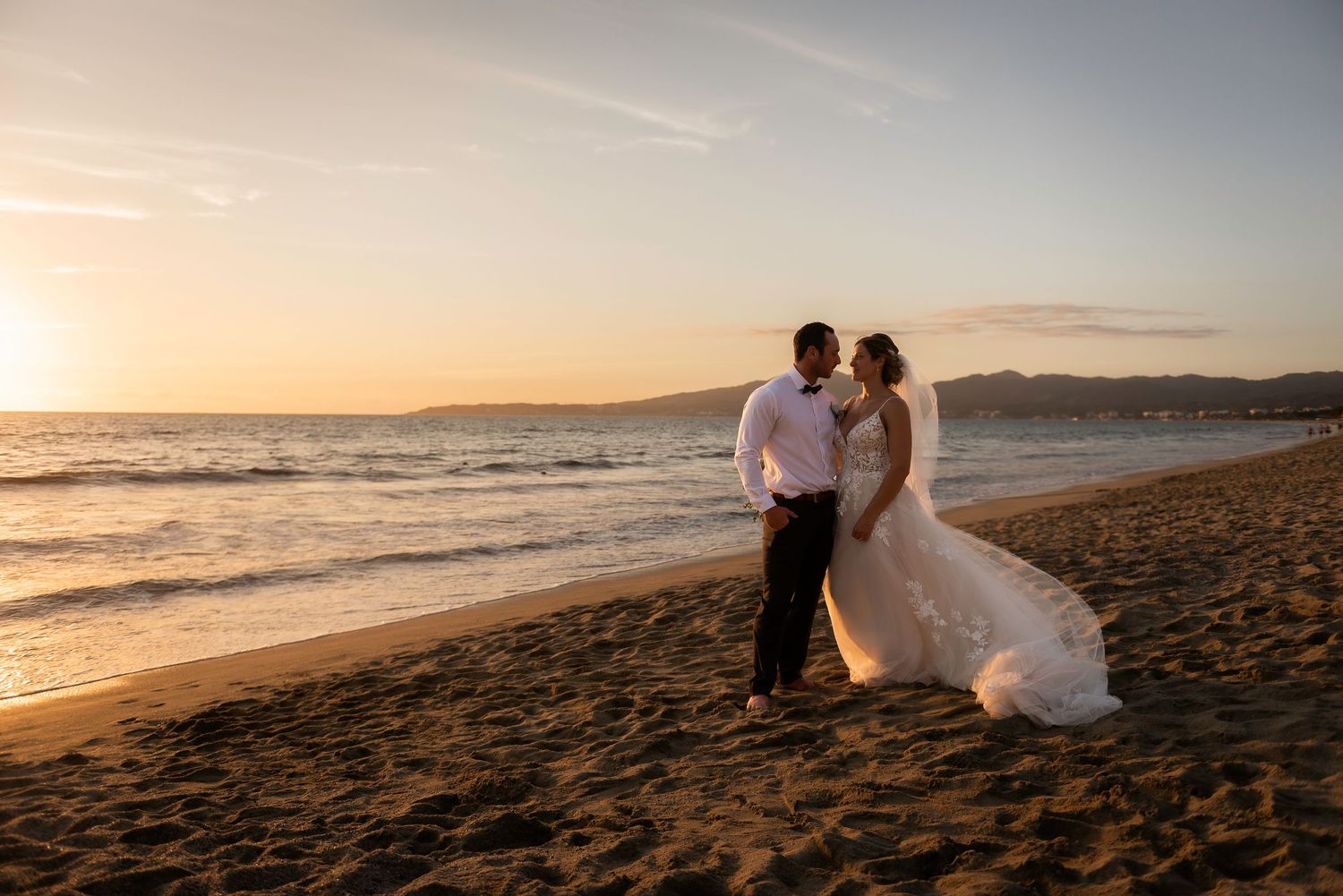 Bride and groom embrace on a sunset beach with waves crashing behind them.