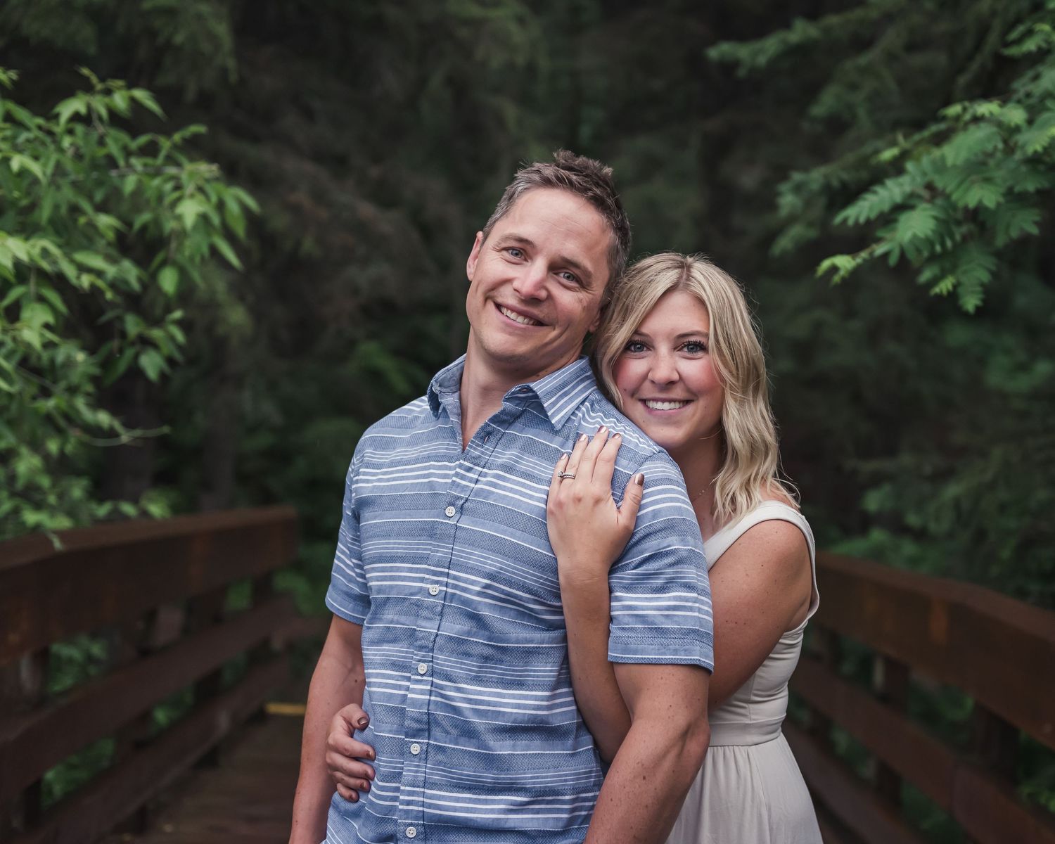 Two people in casual attire embrace on a wooden bridge surrounded by lush green foliage.