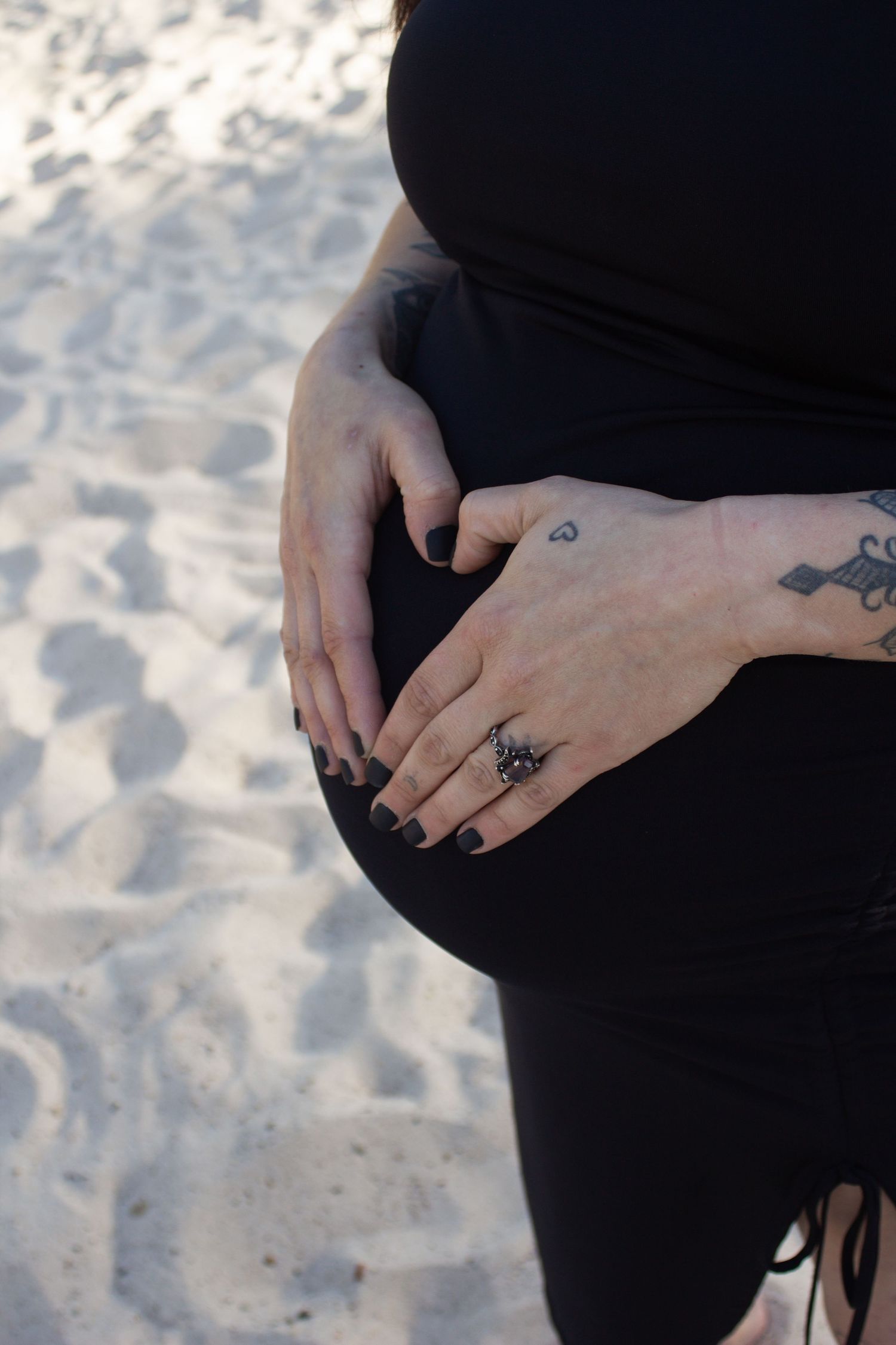 Hands with tattoos rest on a pregnant belly against a sandy beach background.