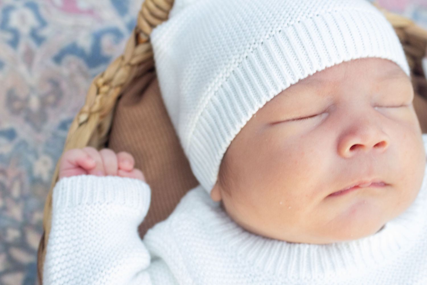 Sleeping baby wearing white knit hat rests in woven basket on floral background.