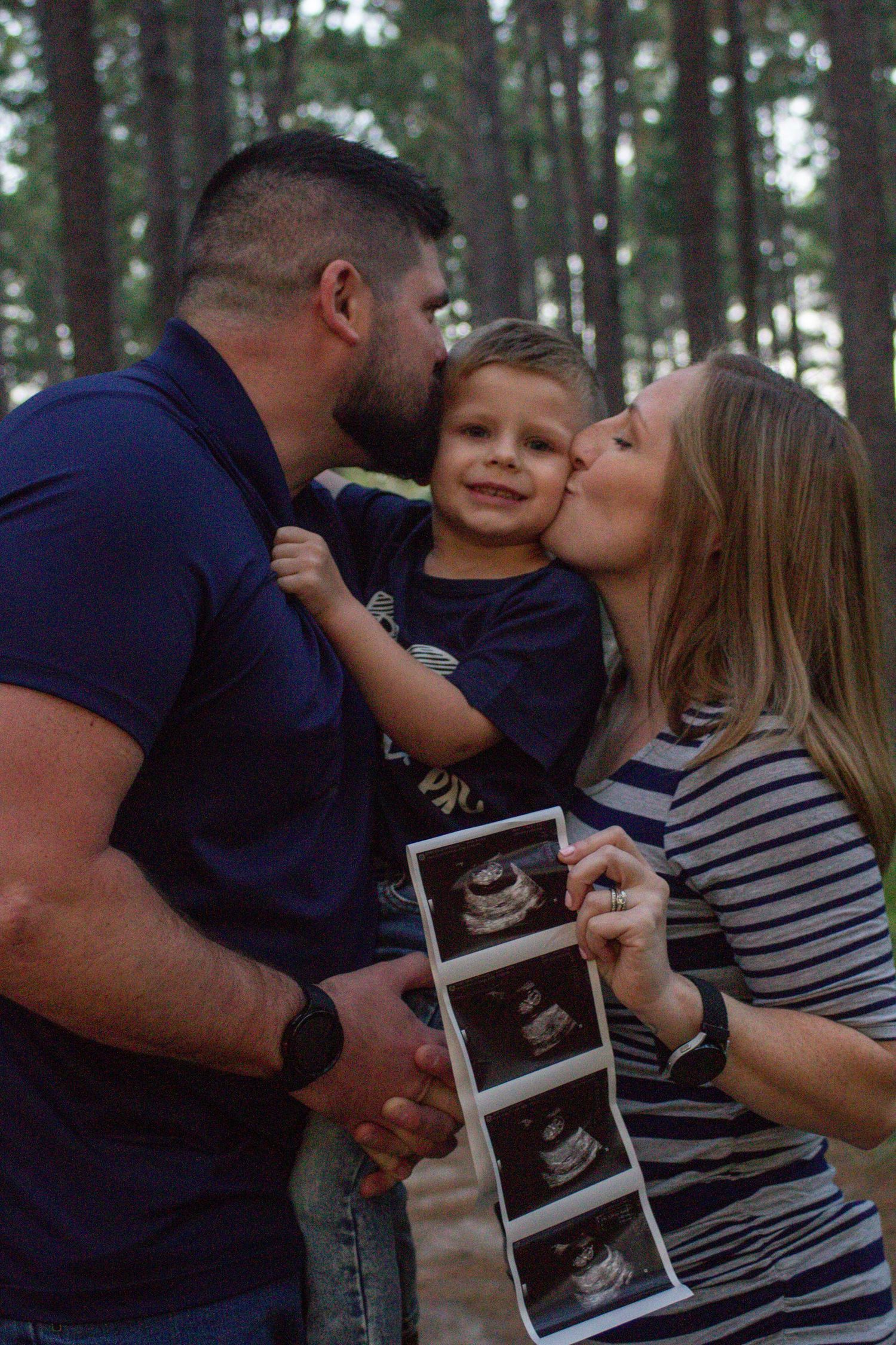 Family shares tender moment in pine forest while holding ultrasound images.