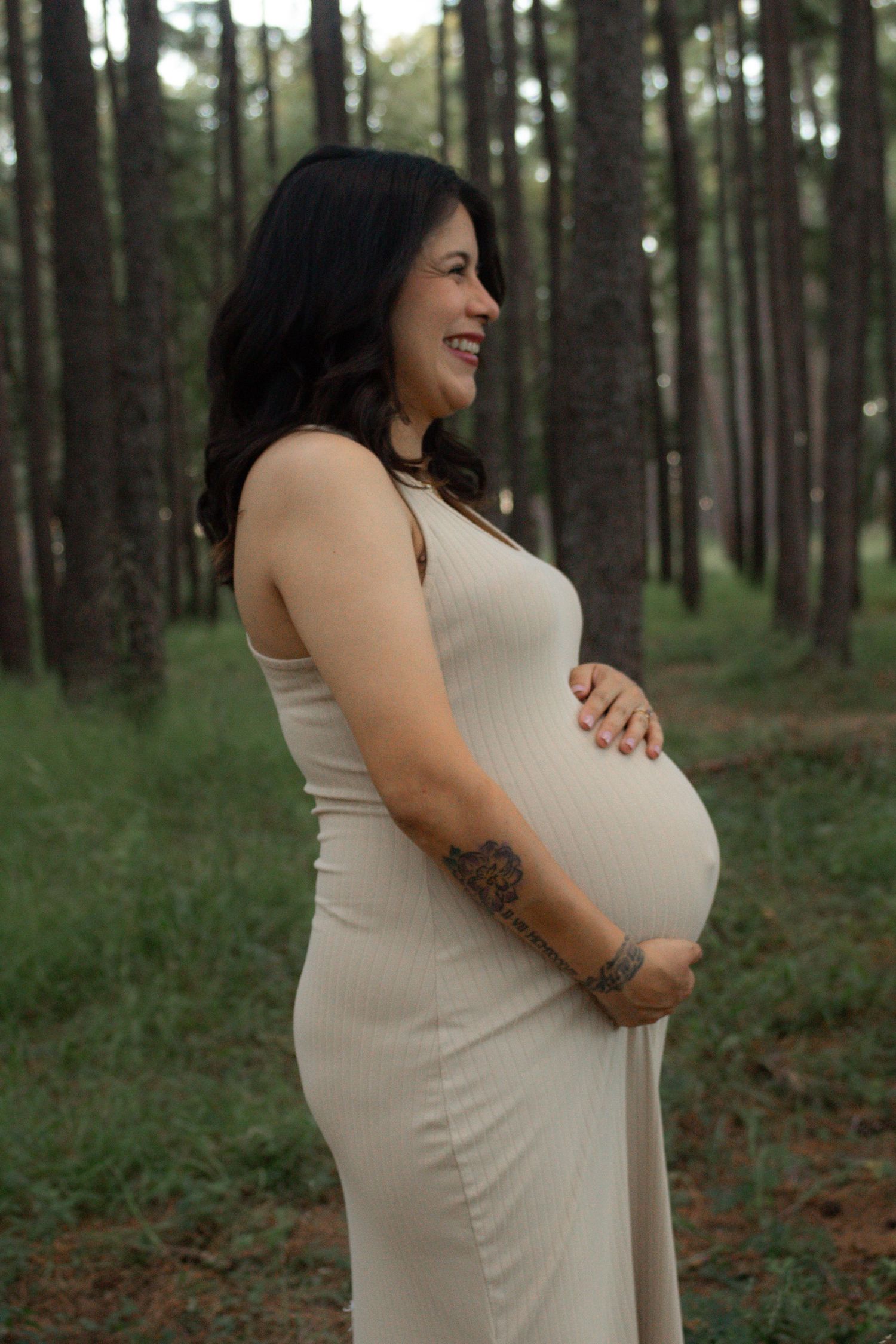 Expectant mother in cream dress stands smiling in forest setting.