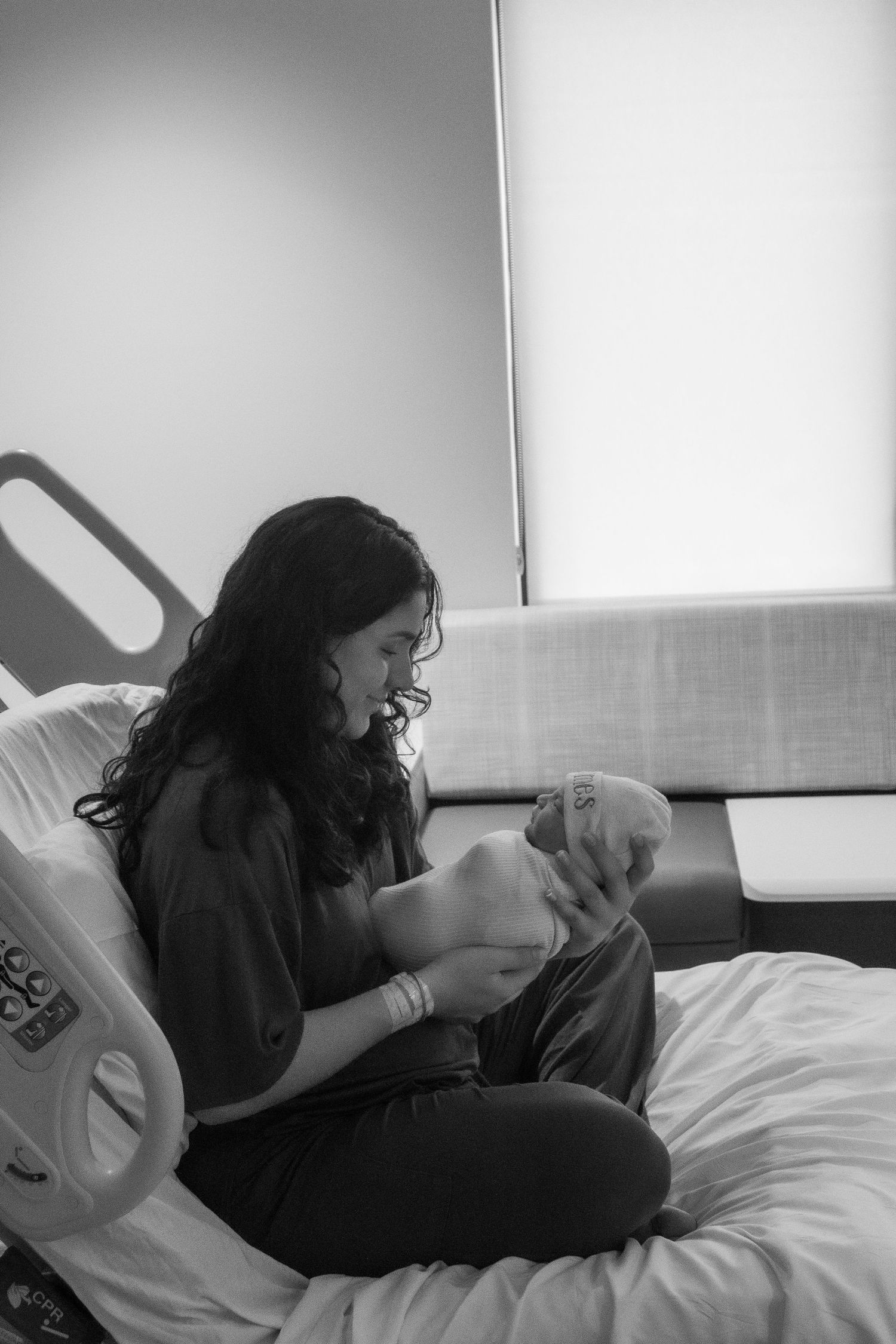 A woman in dark clothing sits on a hospital bed holding a small infant.