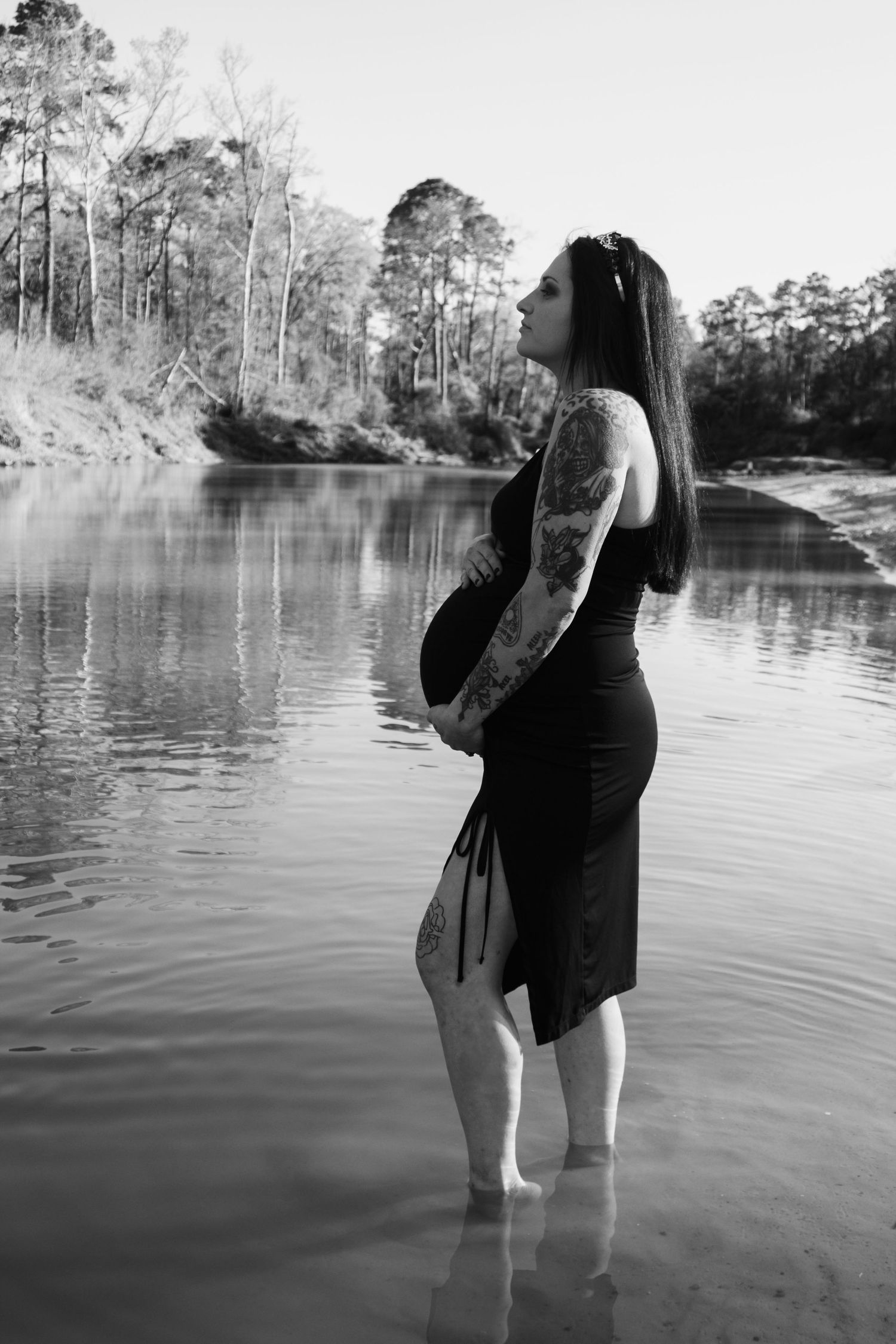 A silhouetted pregnant figure stands ankle-deep in calm water with trees reflecting on the surface.