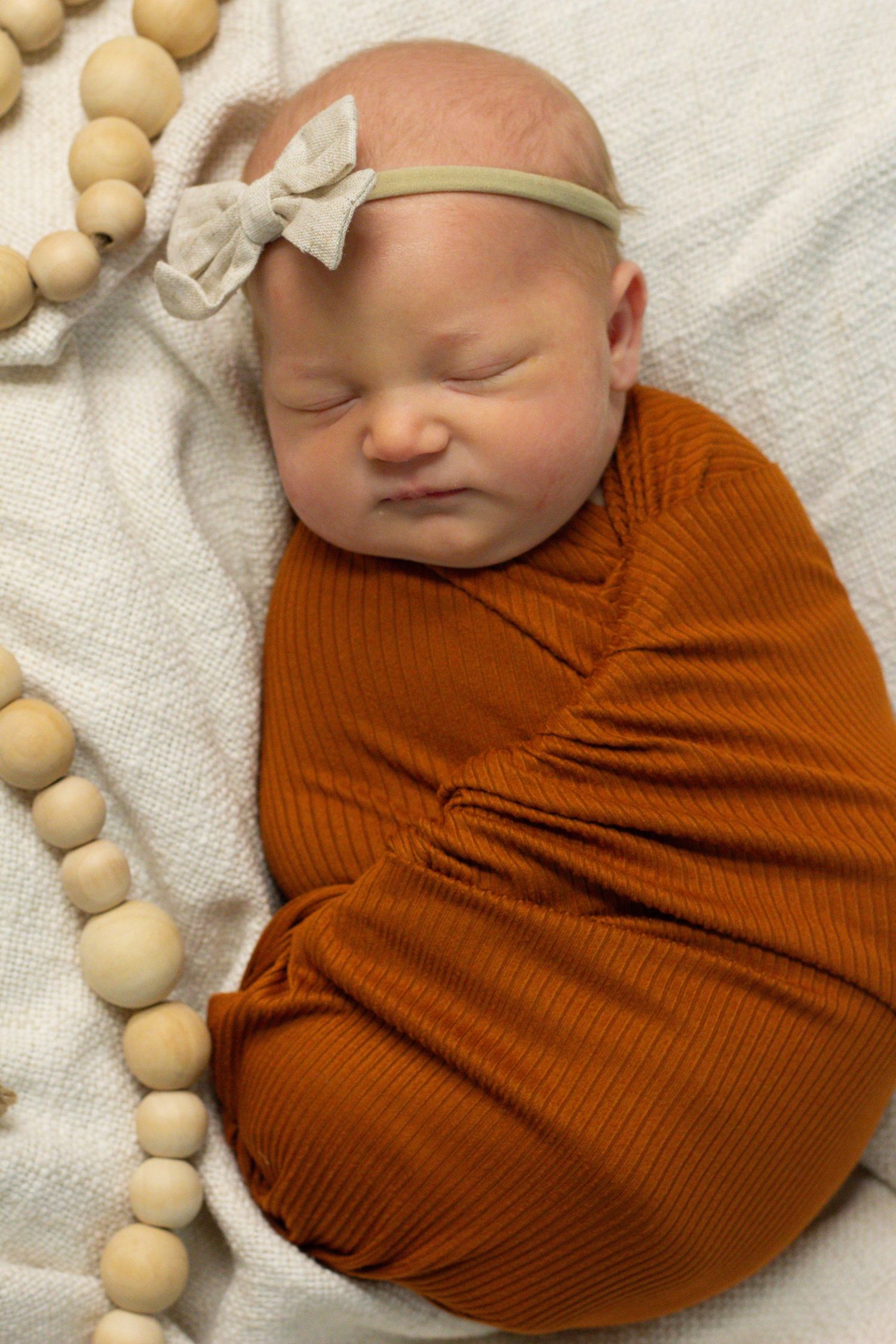 Sleeping newborn in rust colored swaddle with bow headband next to wooden beaded garland.