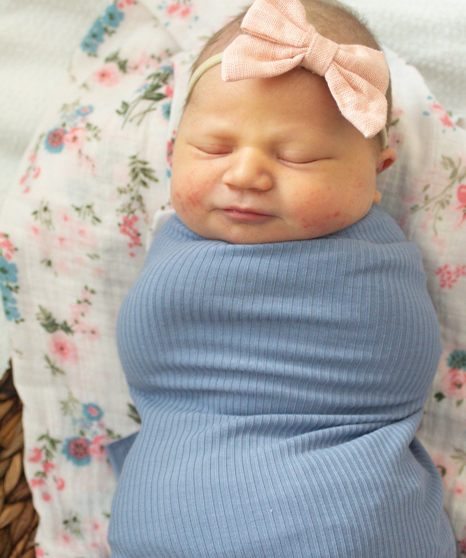 Swaddled newborn in blue wrap with peach bow resting on floral blanket.