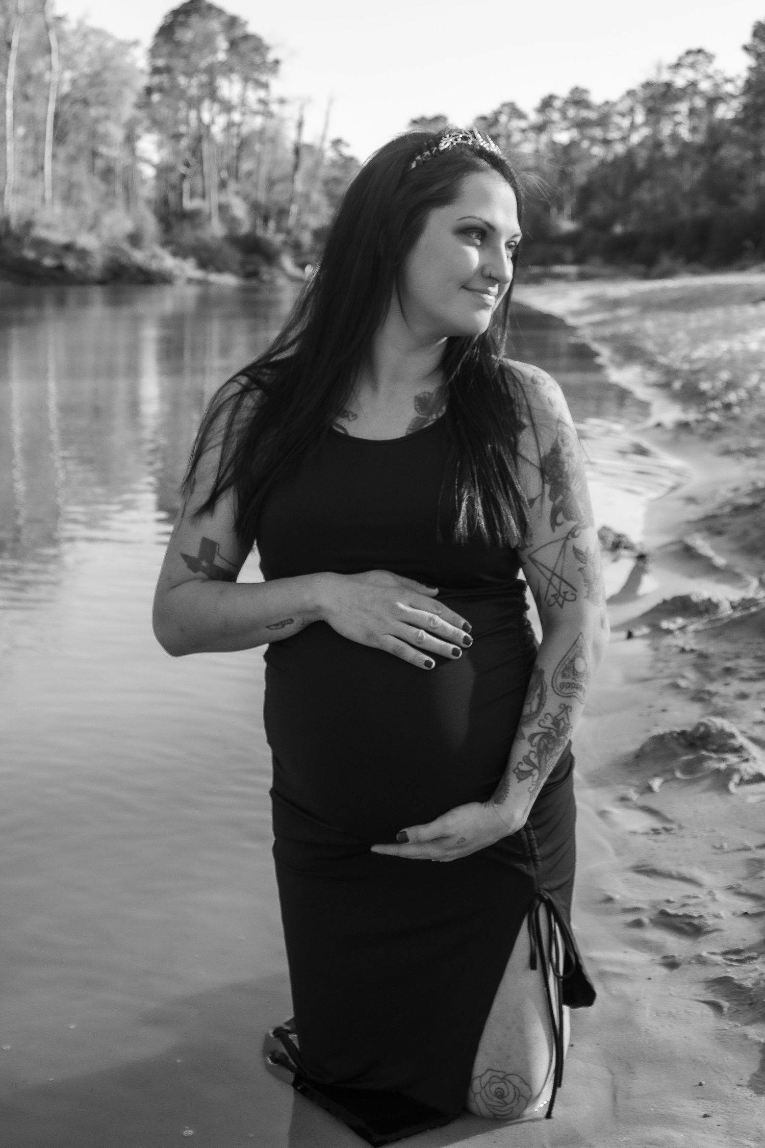 Black and white maternity portrait by a lake with pine trees reflecting on the calm water.