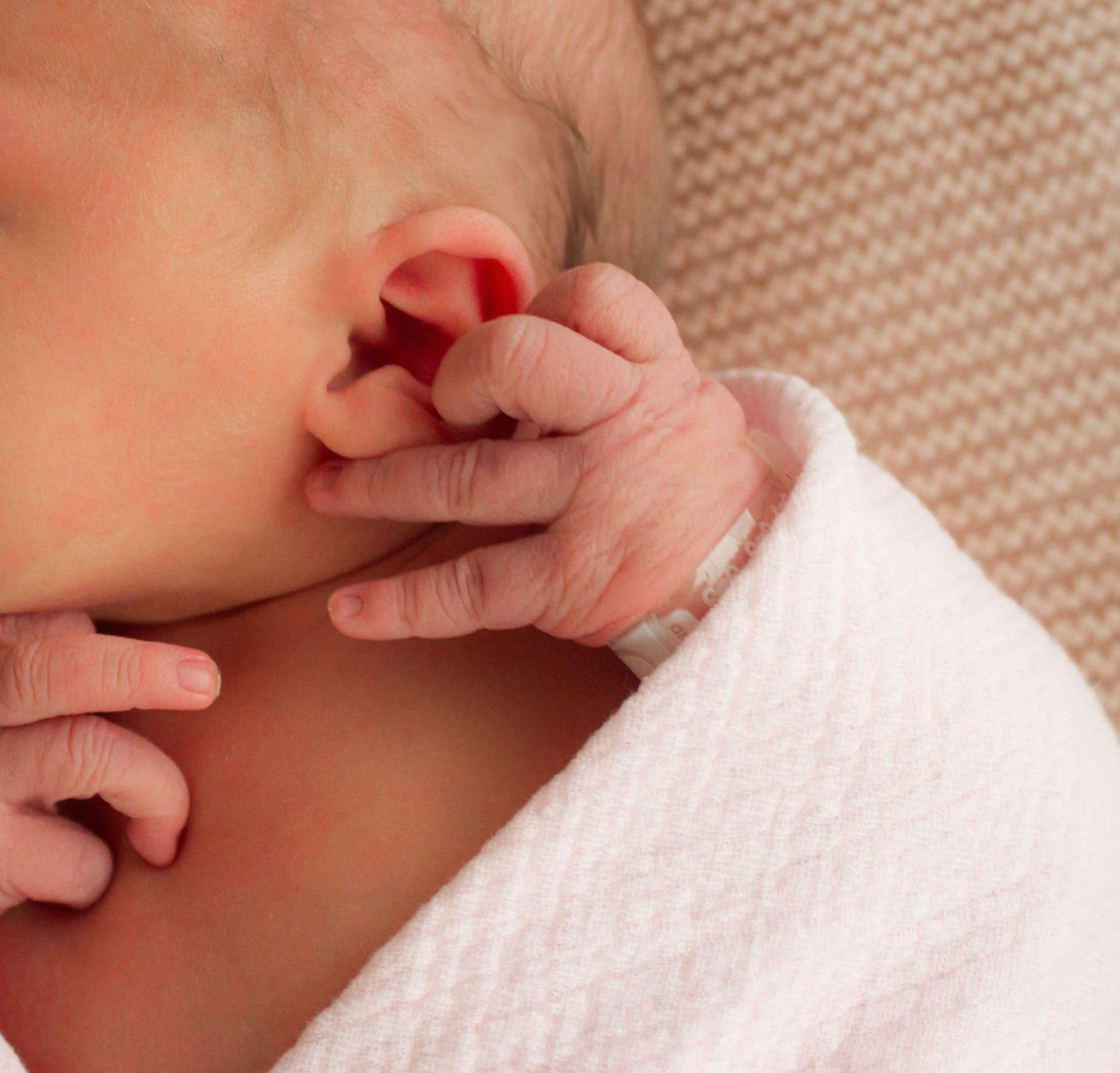 Close-up of sleeping newborn's hand near ear wrapped in white blanket.
