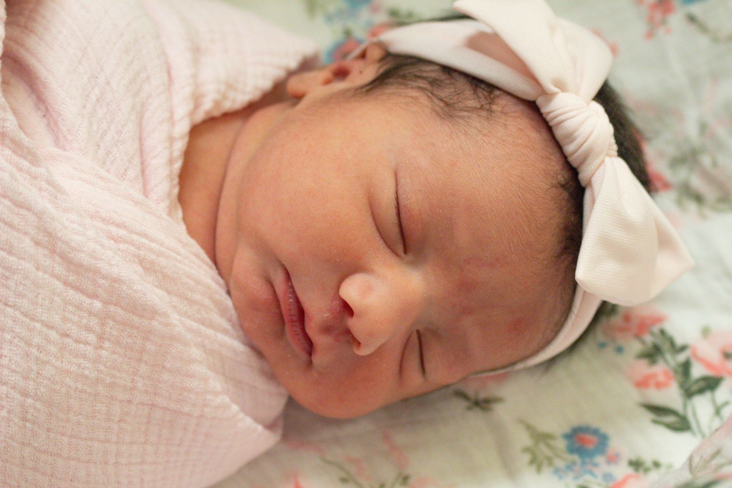 Sleeping newborn wearing pink swaddle and white bow headband on floral pattern fabric.