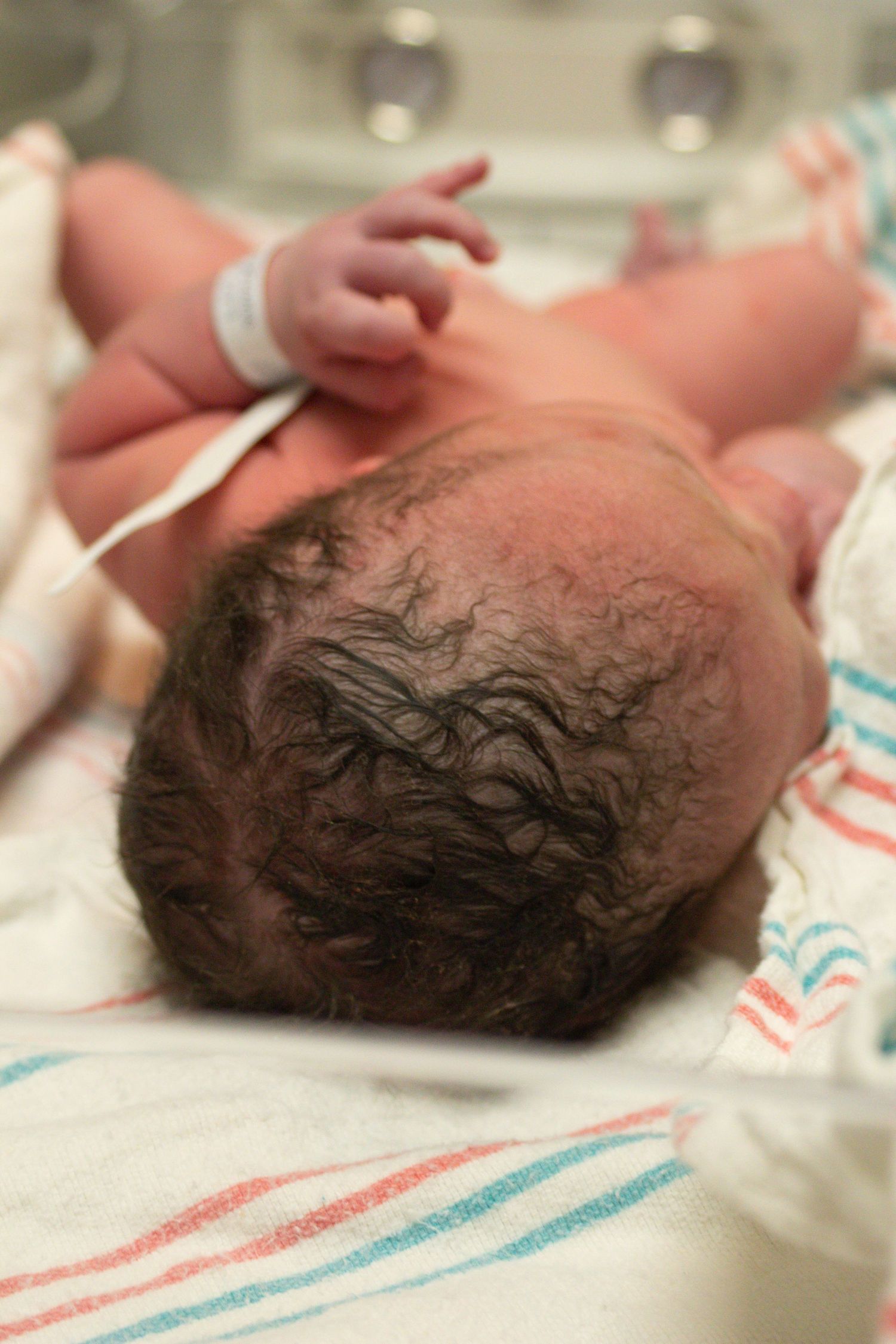 Newborn with dark curly hair lying on striped hospital blanket.