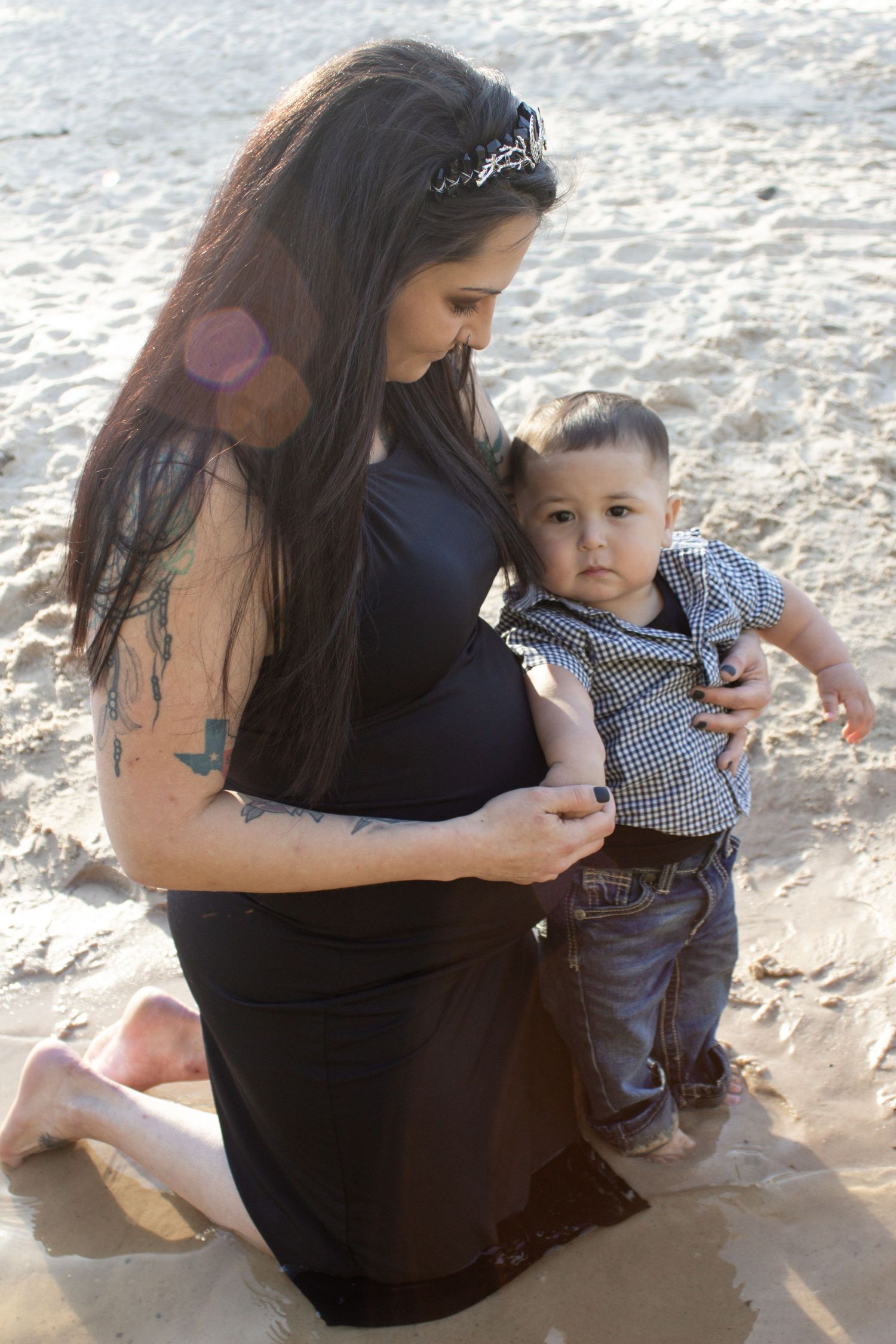 A mother in a dark dress kneeling on a beach with her young child during sunset.