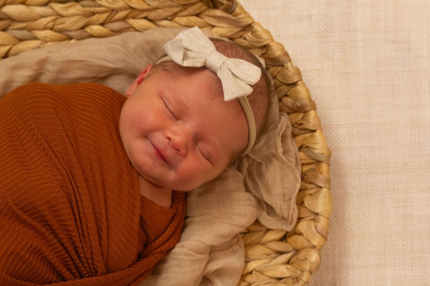 A newborn baby swaddled in a rust-colored wrap with a white bow sleeping peacefully in a woven basket.