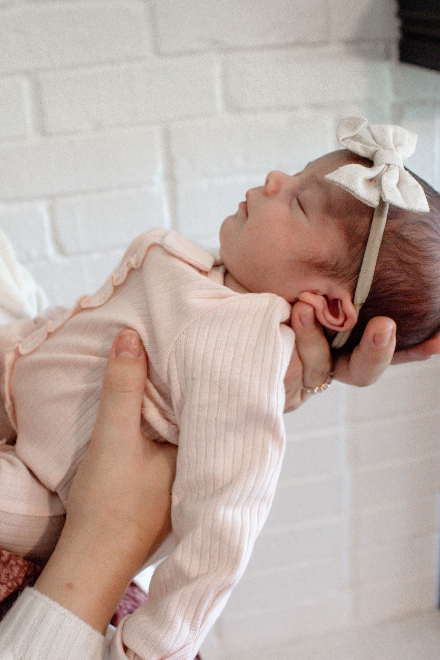 A sleeping newborn in pale pink clothing with white bow against white brick wall.