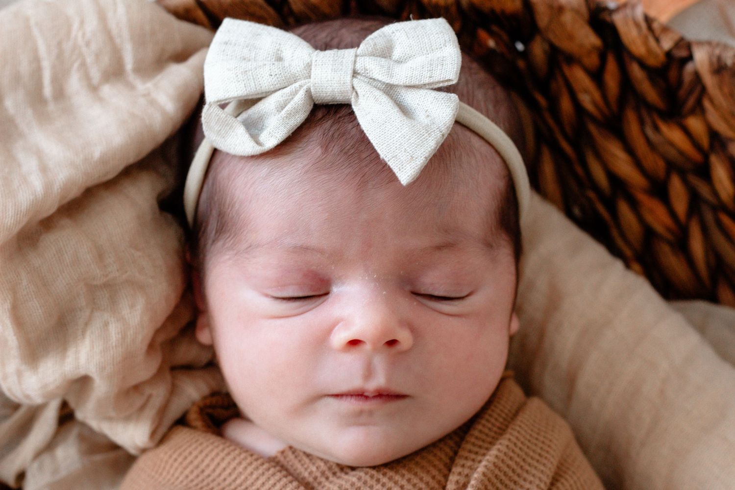 A sleeping newborn wearing a white bow headband against neutral toned bedding.
