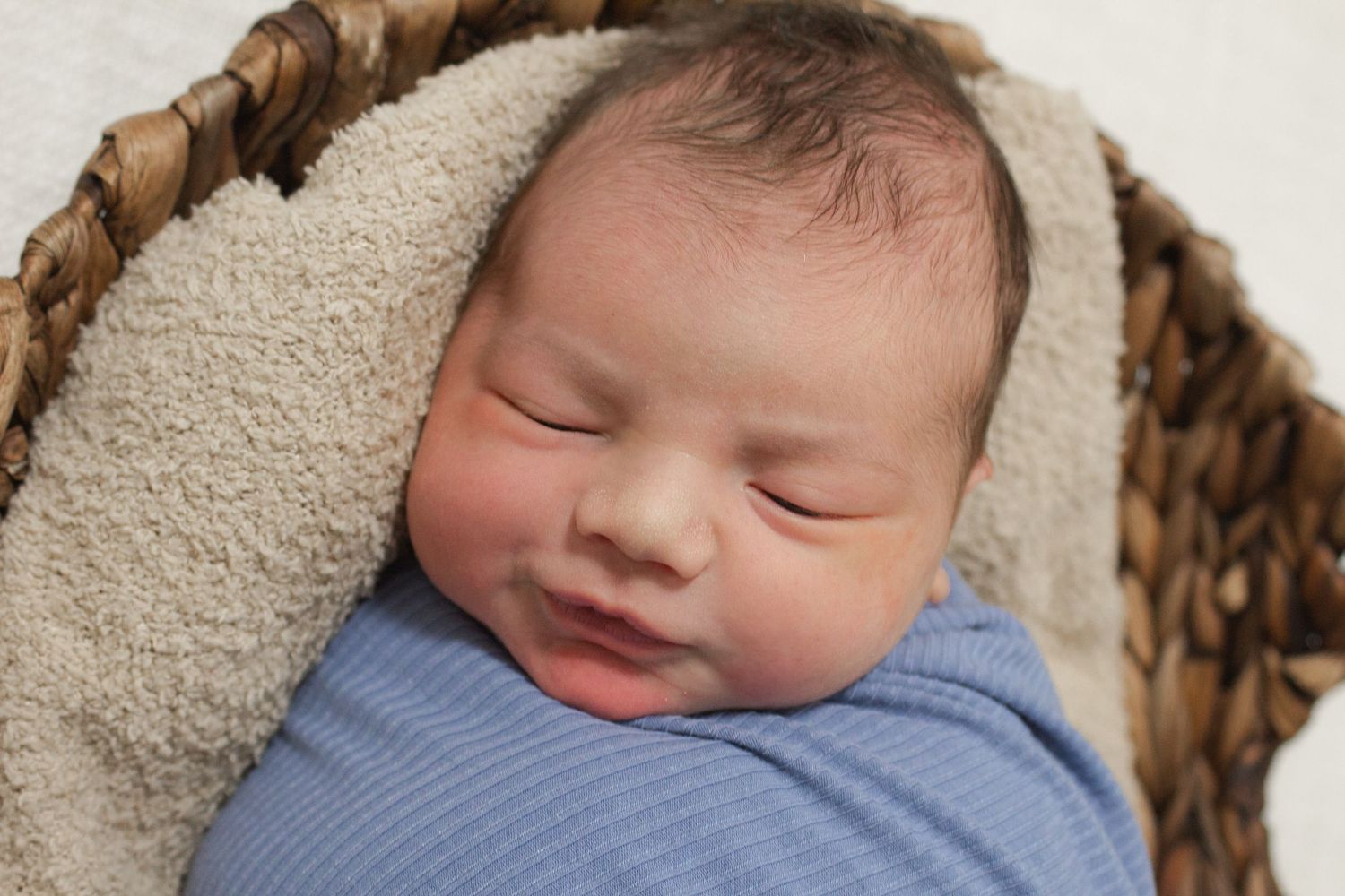A sleeping newborn baby swaddled in blue fabric resting in a woven basket.