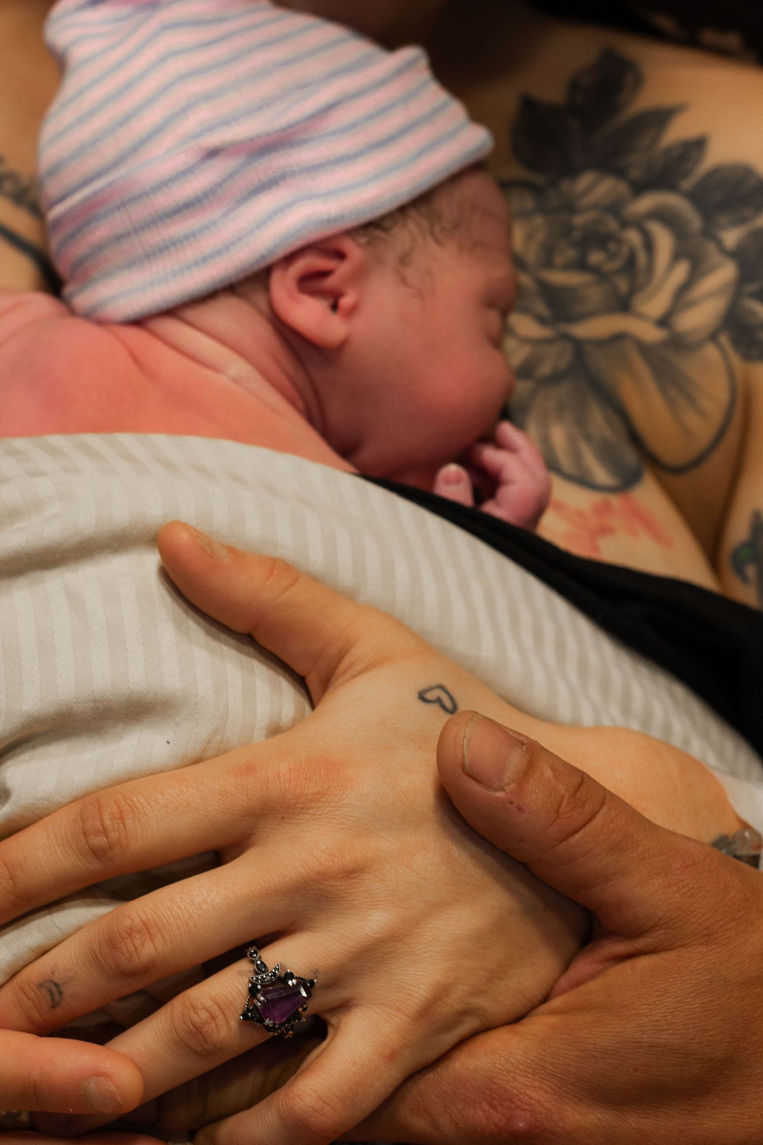 Newborn baby wearing striped hat rests against parent with floral tattoo and ring.