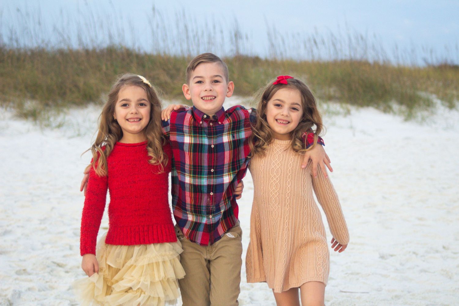 Three children standing together on a sandy beach with dunes in the background.