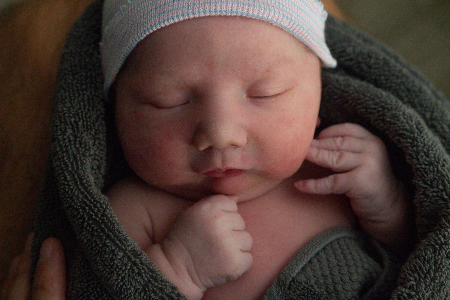 Peaceful newborn in grey knit cap and wrap sleeping with tiny hands near face.