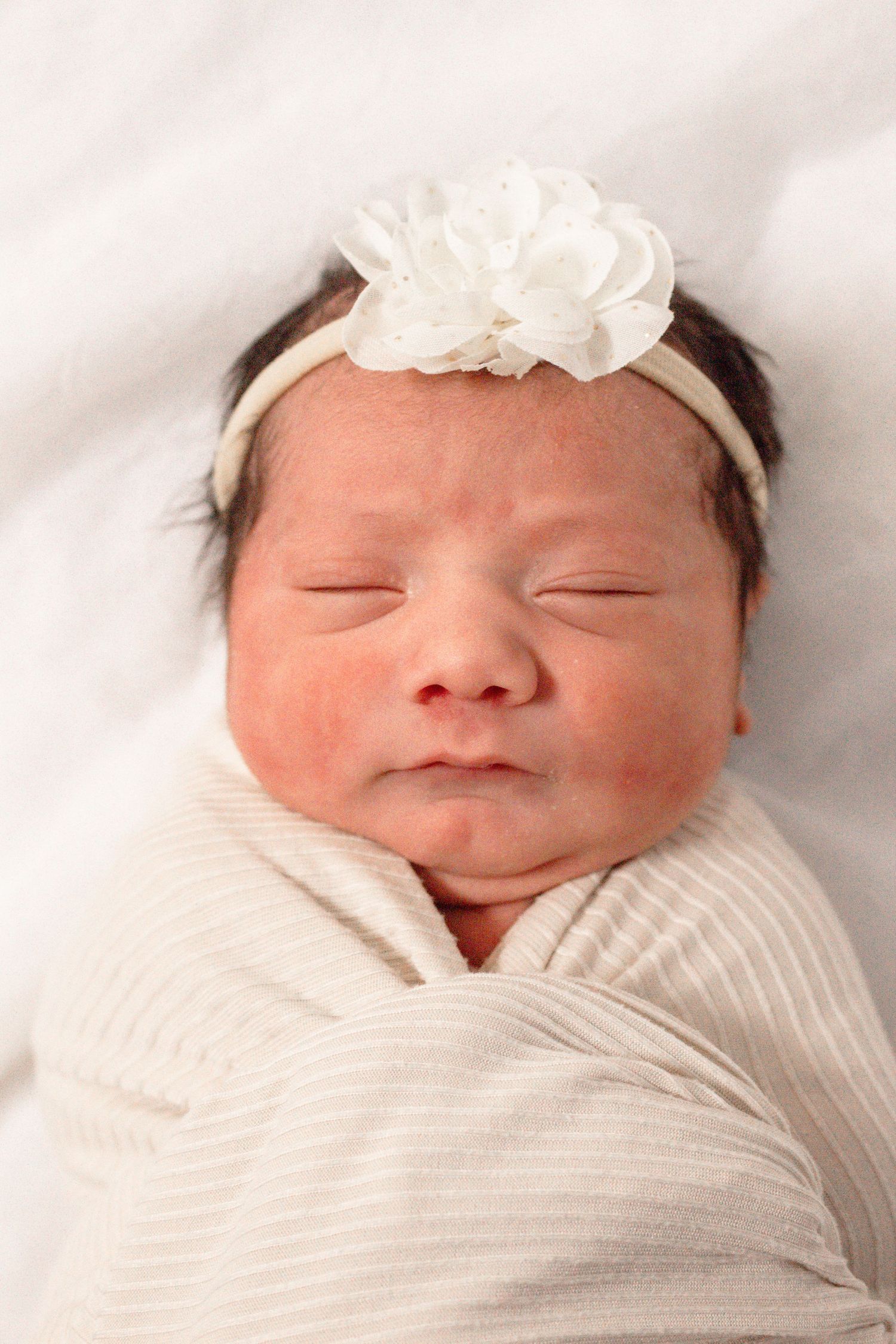 Peaceful sleeping newborn wrapped in white swaddle wearing delicate white flower headband.