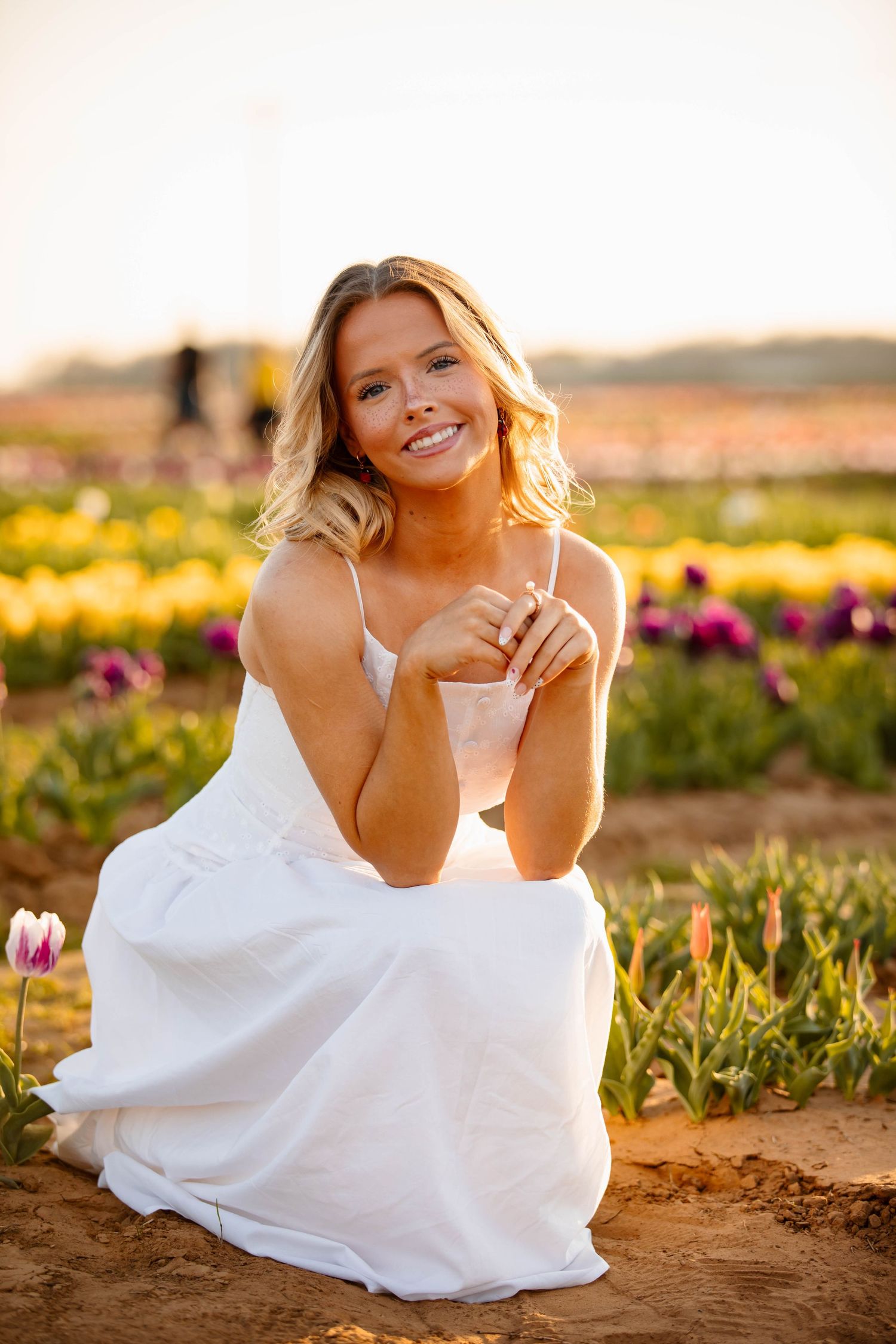 A person in a flowing white dress lies on the ground at sunset in a flower field during a photo session.