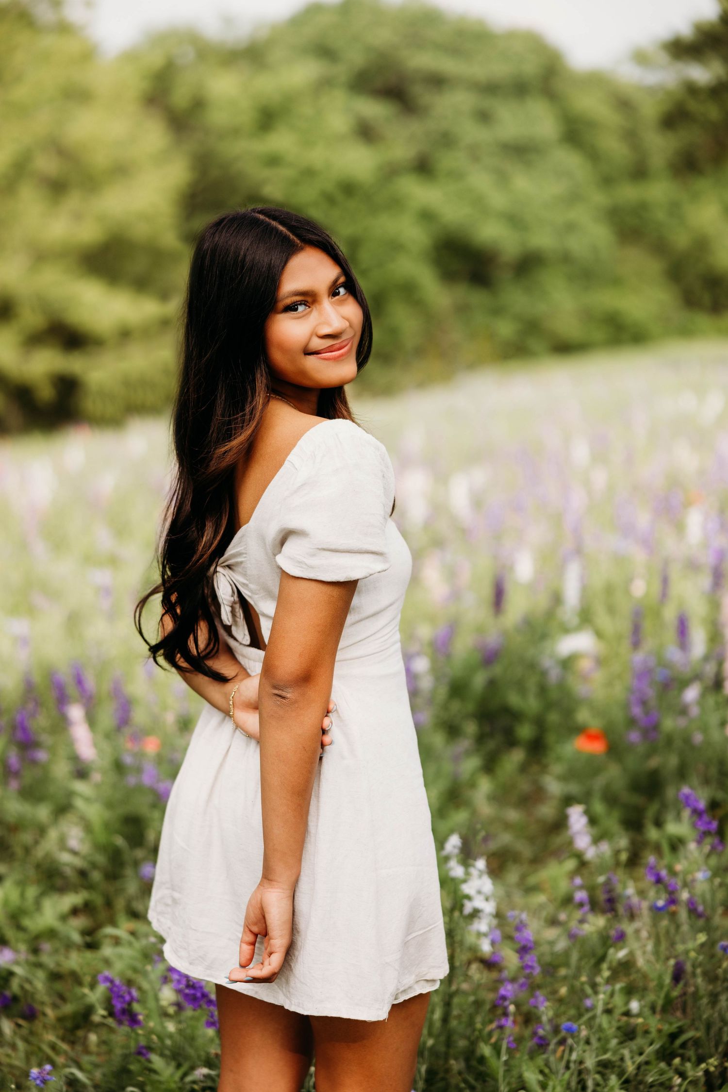 Young woman in white dress smiling over shoulder in vibrant wildflower field with purple blooms and green trees.