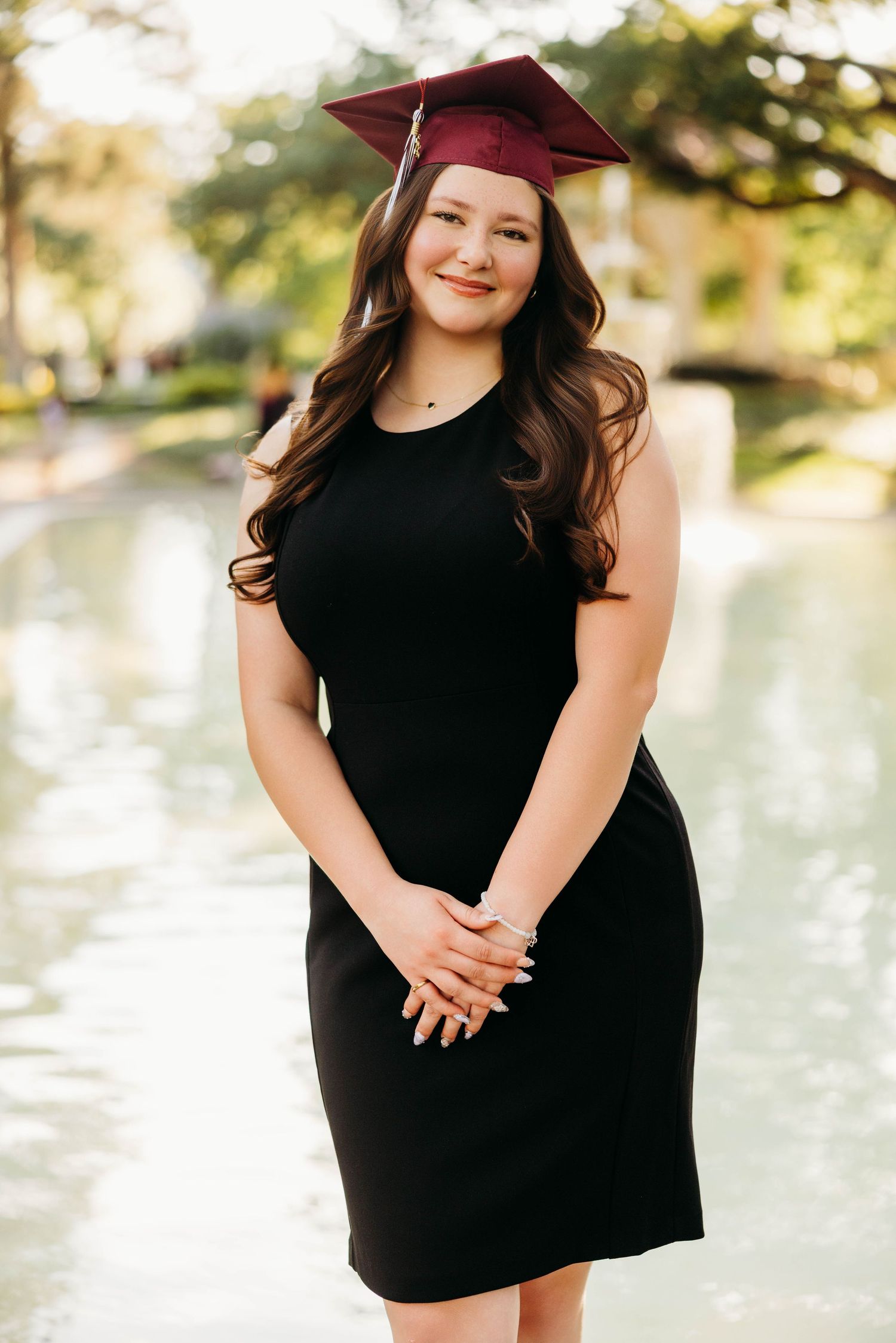 Young woman in black dress and maroon graduation cap smiling outdoors near a reflective pond.