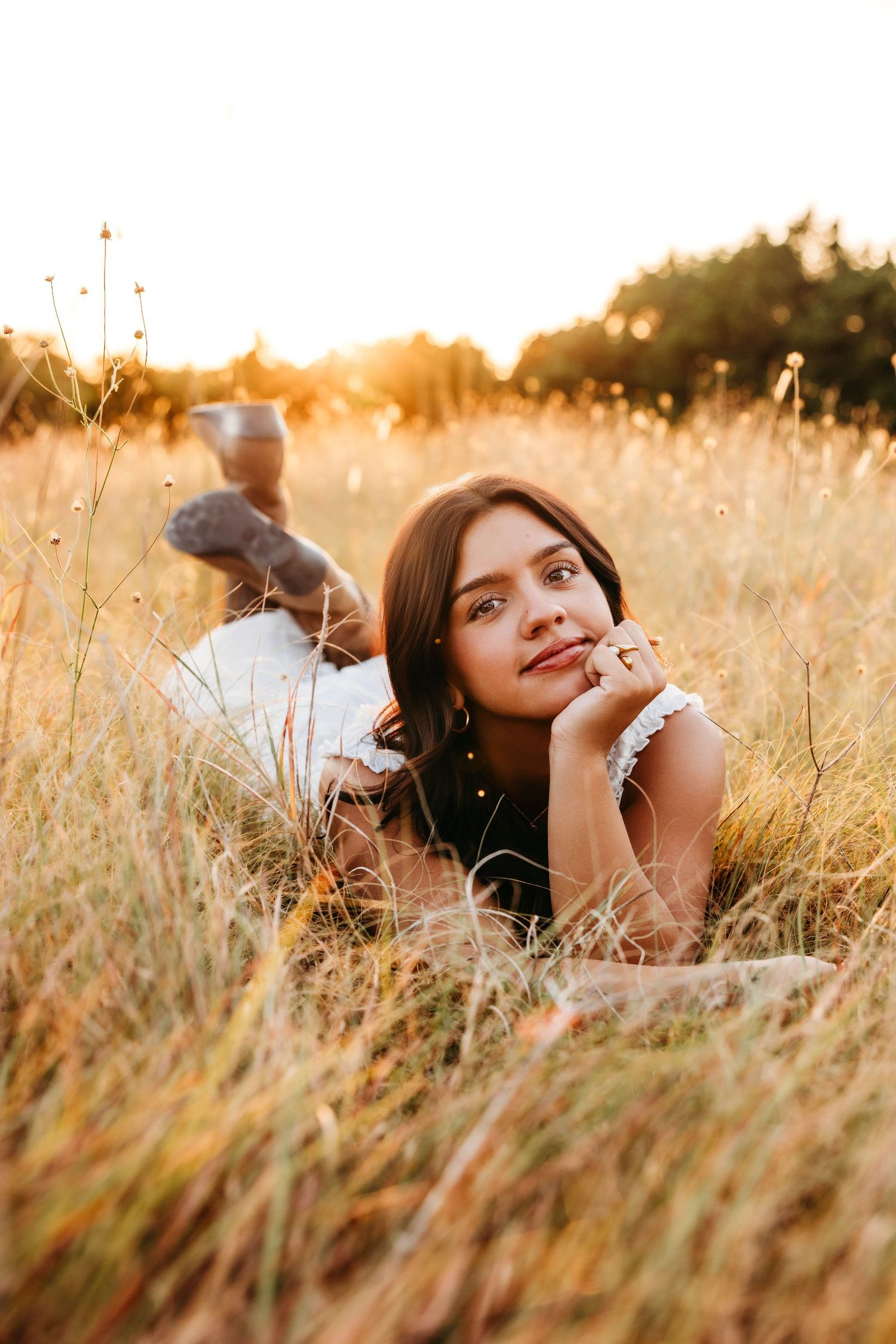 Young woman lying in golden grass field at sunset, resting chin on hand, wearing white dress, with warm backlit glow.