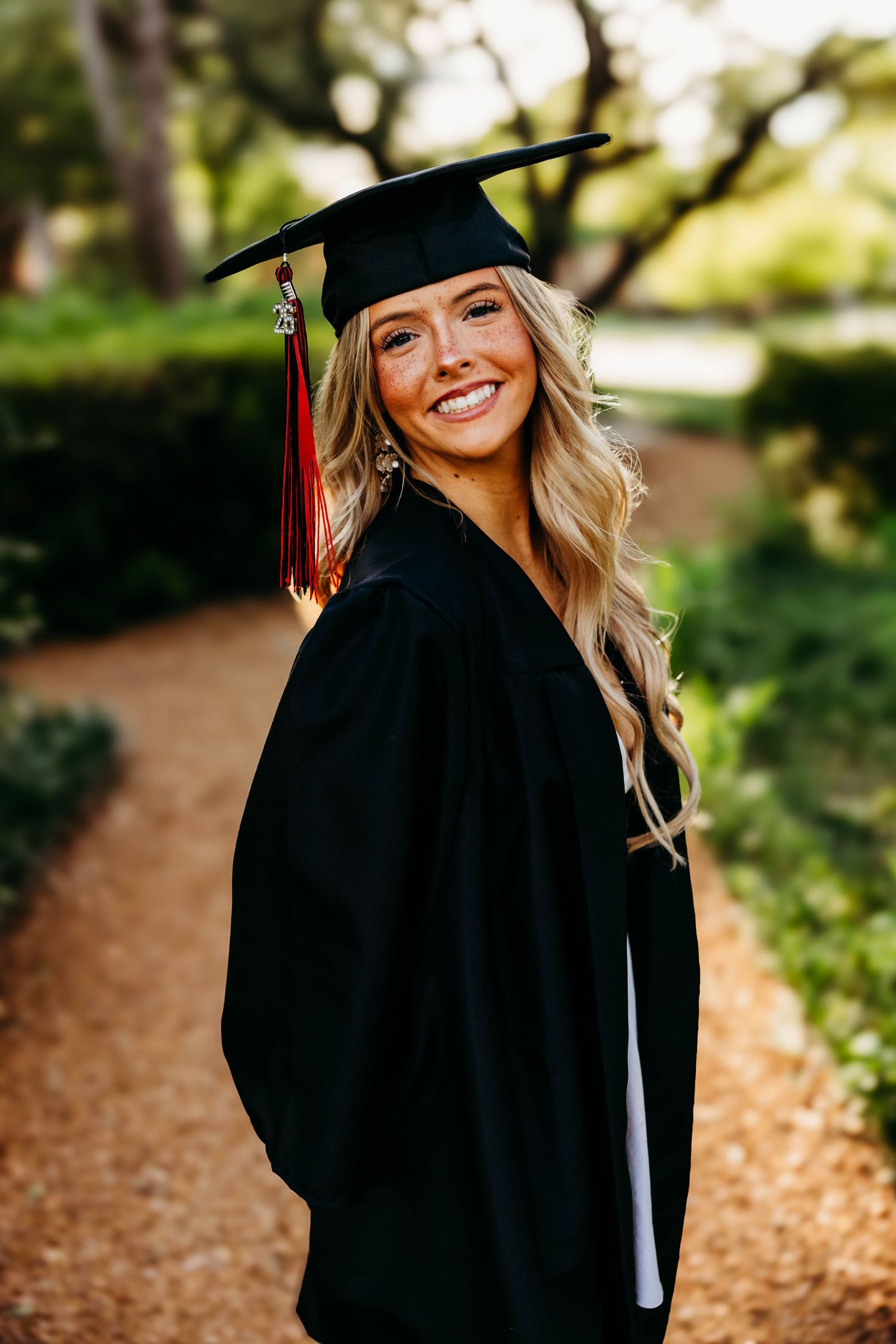 Smiling graduate in black cap and gown with red tassel poses outdoors on a tree-lined path.