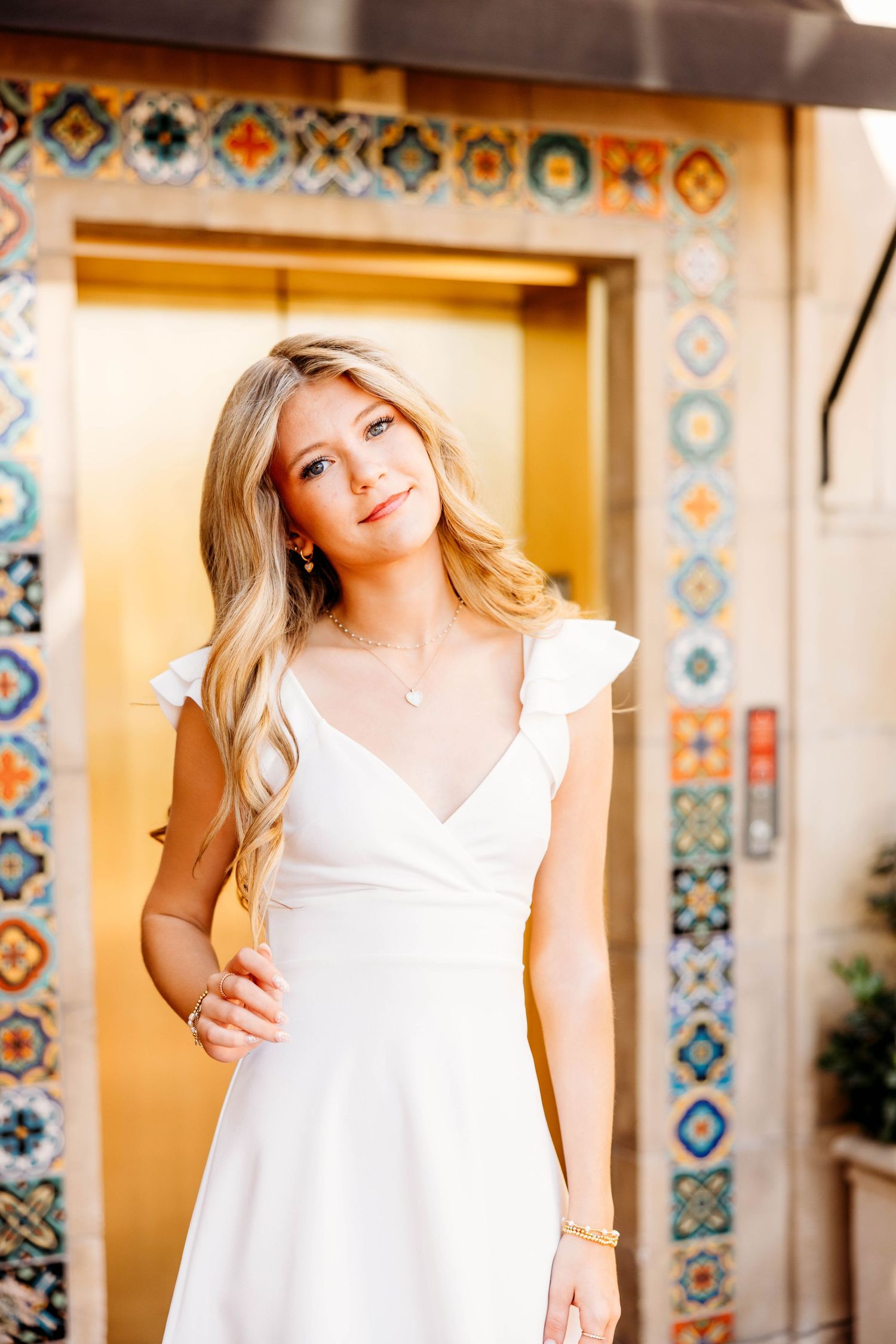 Woman in white dress poses against decorative tiled wall with geometric patterns in warm lighting.