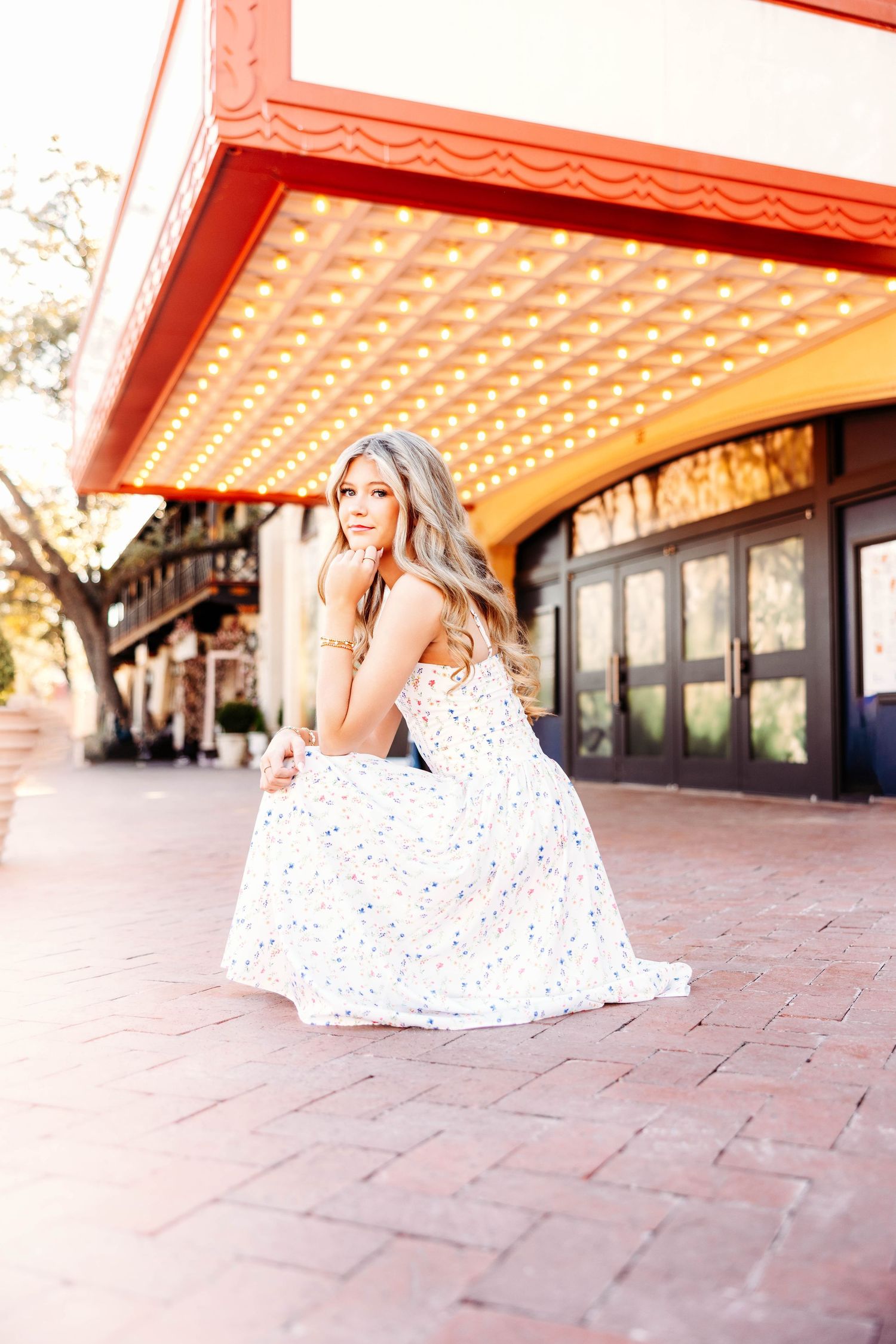 A person in a white polka dot dress sits gracefully on terracotta tiles under a covered walkway with ornate lighting.