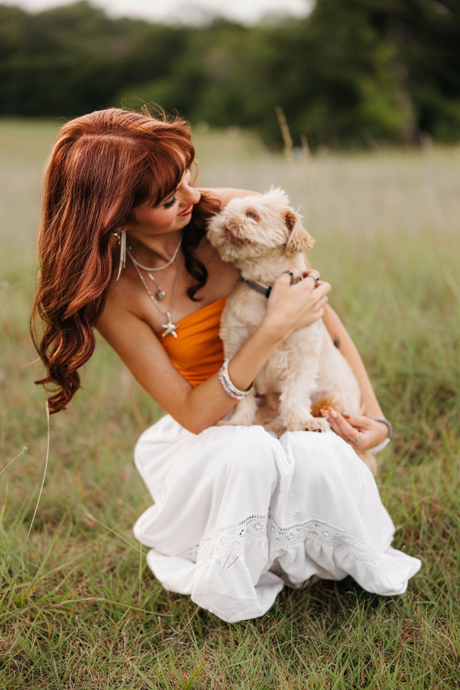A redhead woman in orange top and white skirt crouches in a field, affectionately nuzzling a small fluffy dog.