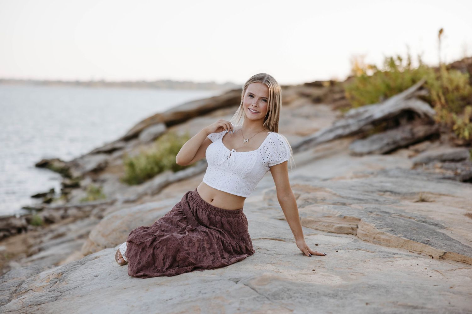 Young blonde woman in white crop top and brown skirt sitting on rocky shoreline smiling outdoors.