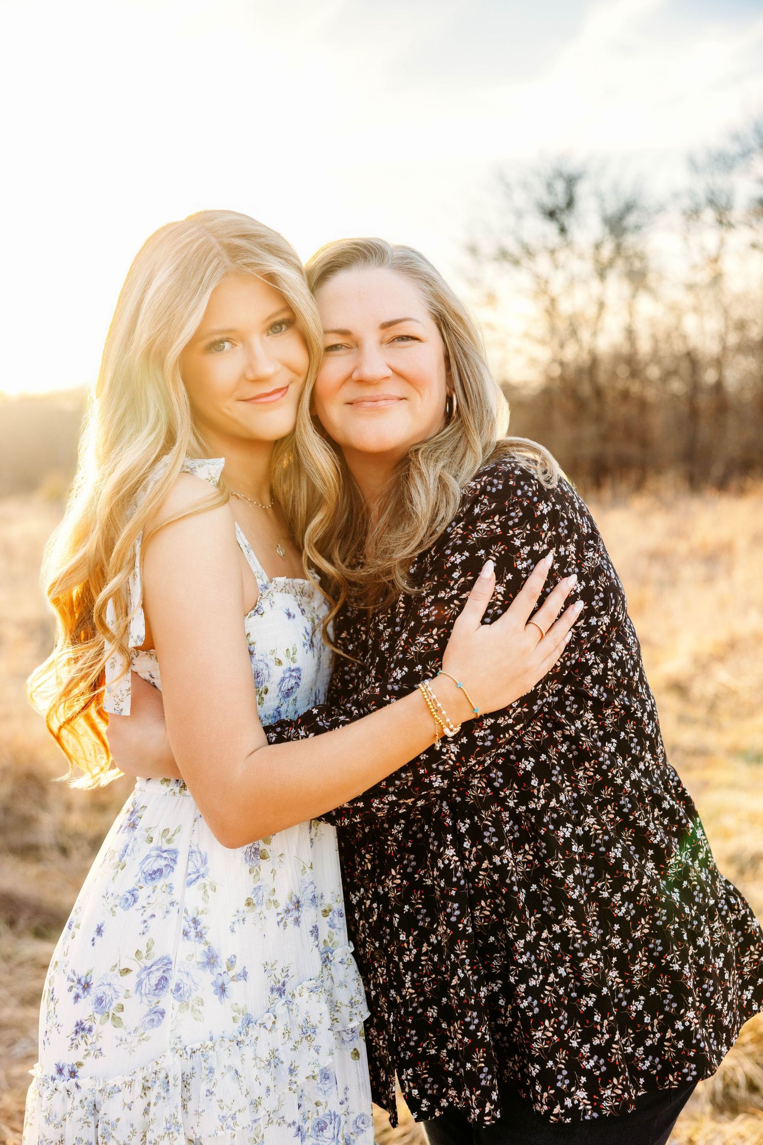 Two people in floral dresses embrace and laugh together in a sunlit field during golden hour.