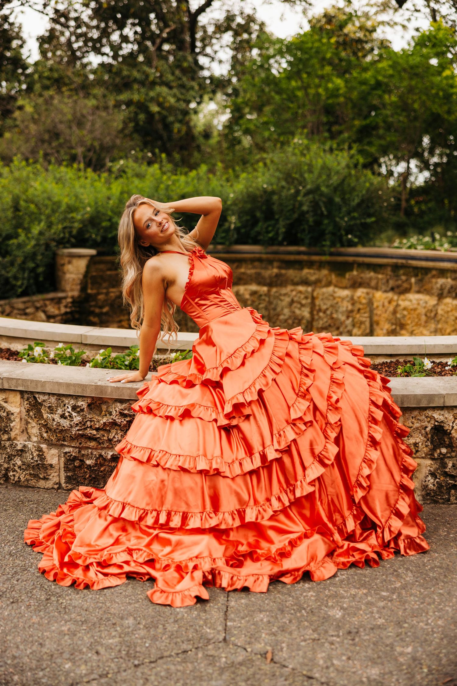 Woman in orange ruffled ballgown posing gracefully in an outdoor garden setting with stone walls and lush greenery.