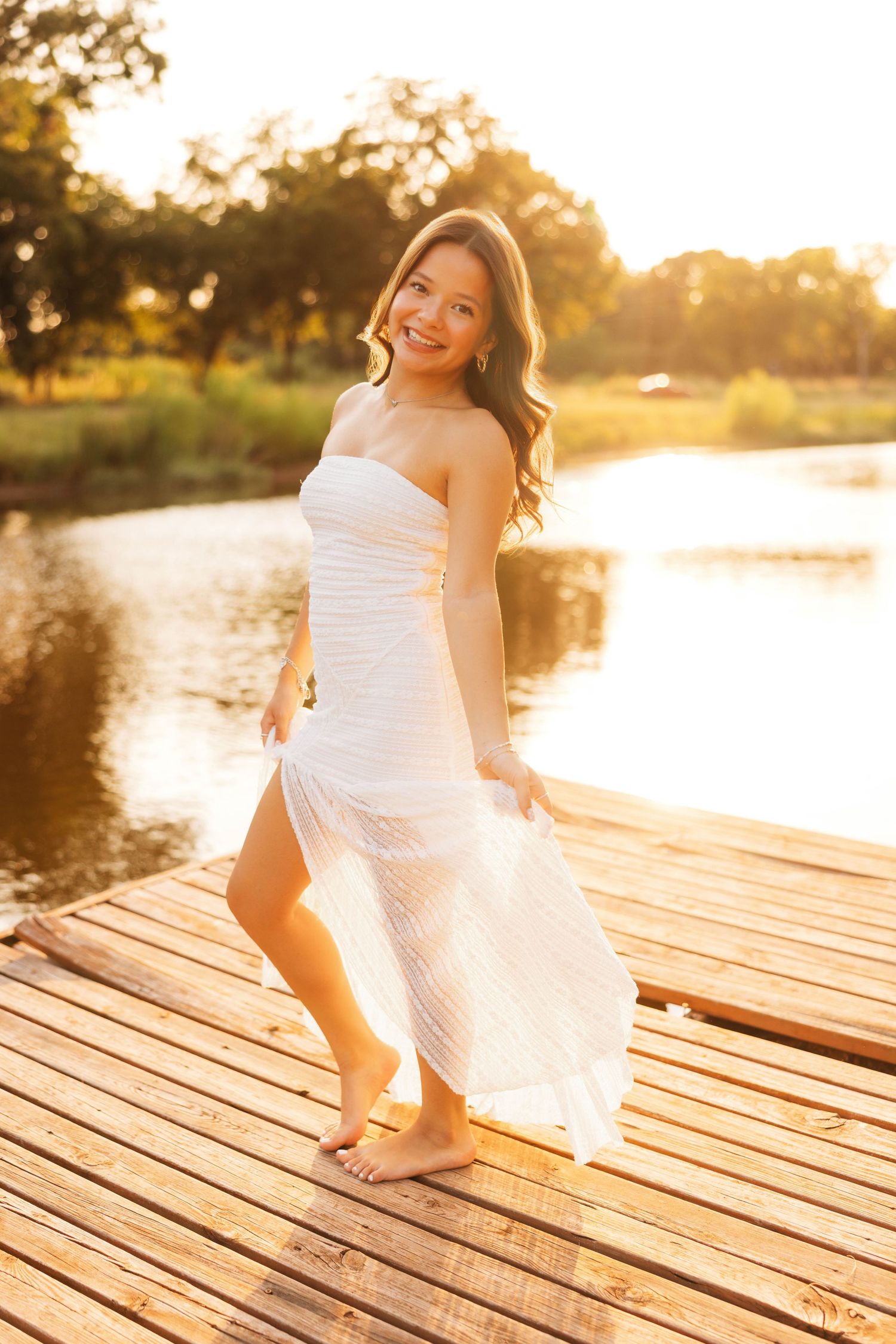Young woman in white strapless dress standing barefoot on wooden dock at golden sunset by a lake.